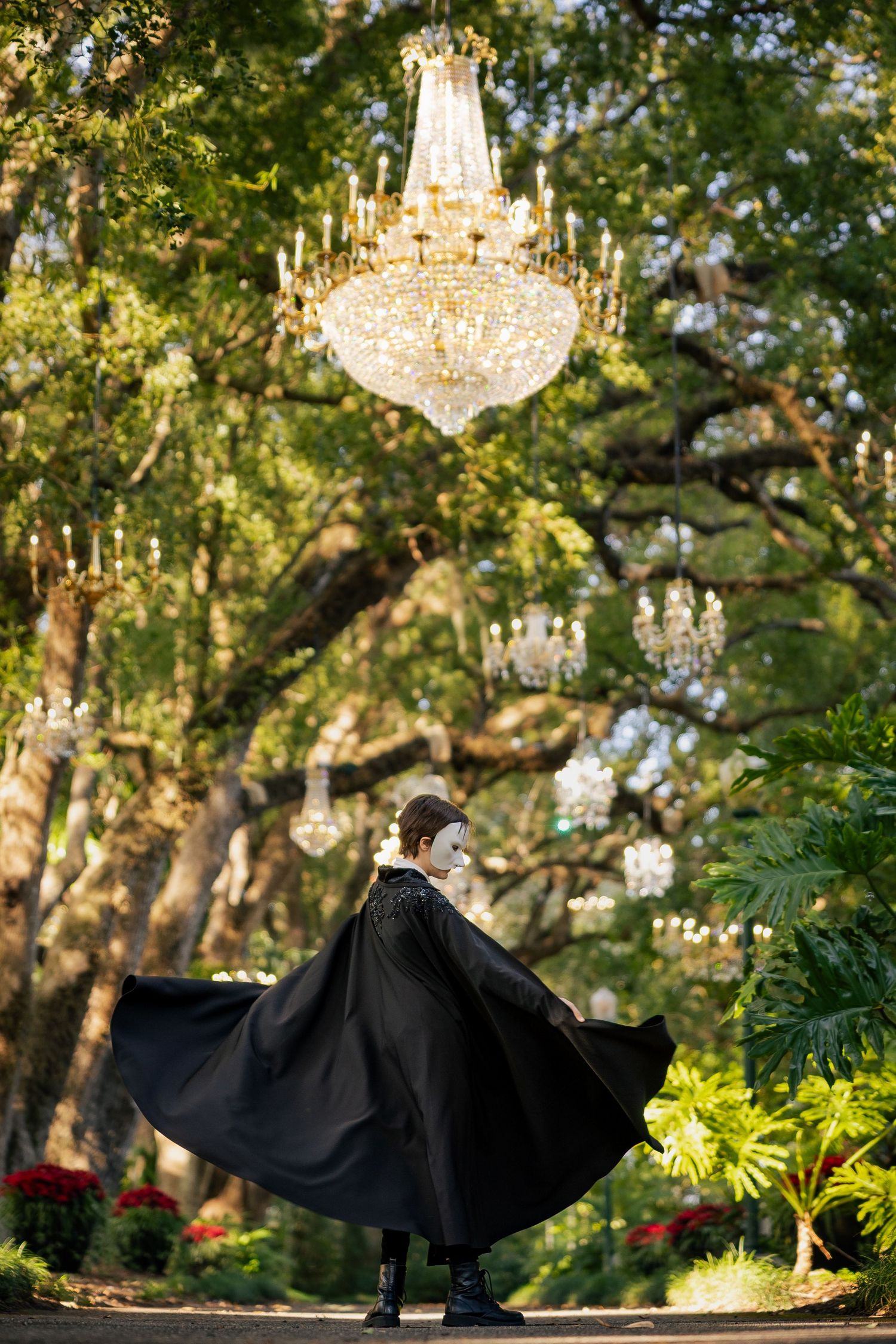 Person in black cape and white mask poses under crystal chandeliers hanging from oak trees in lush garden.