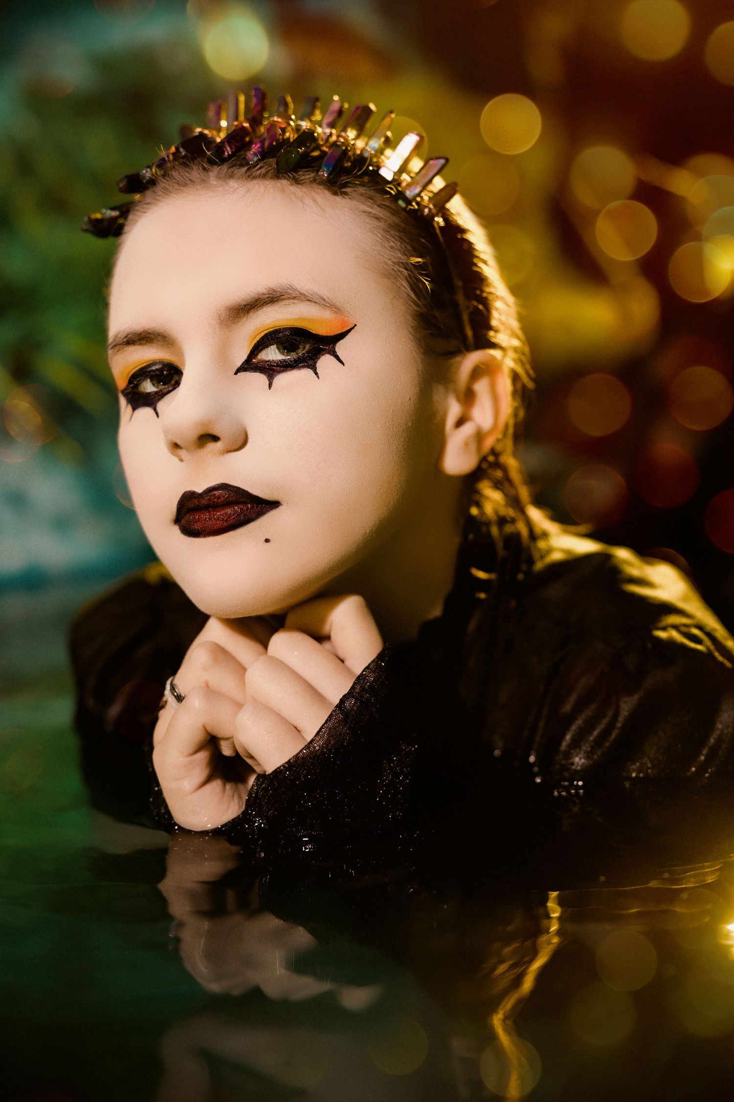 Woman with dramatic gold and black theatrical makeup wearing crystal crown in dark water.