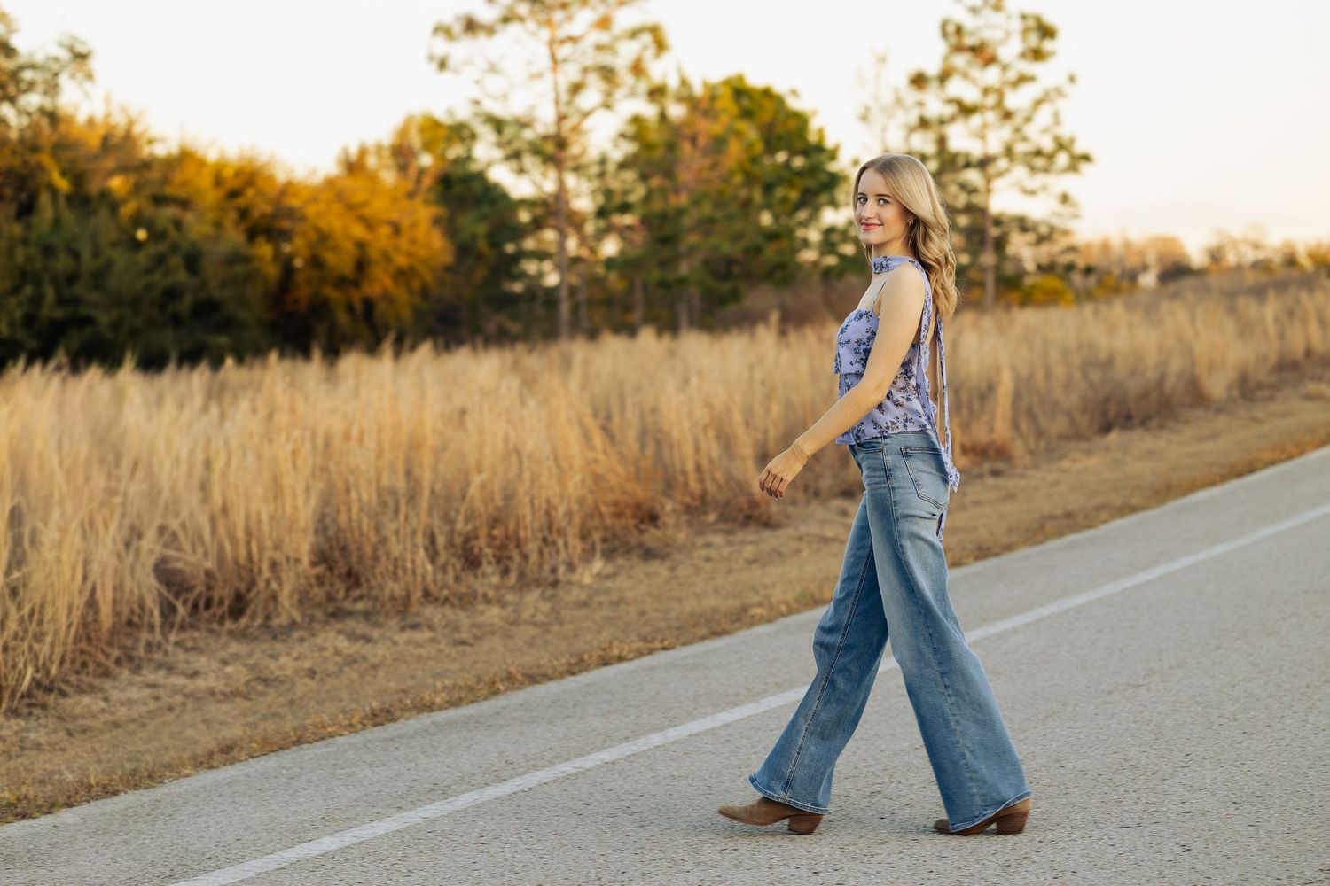 A young woman in wide-leg jeans and floral top walks along a rural road at golden hour.