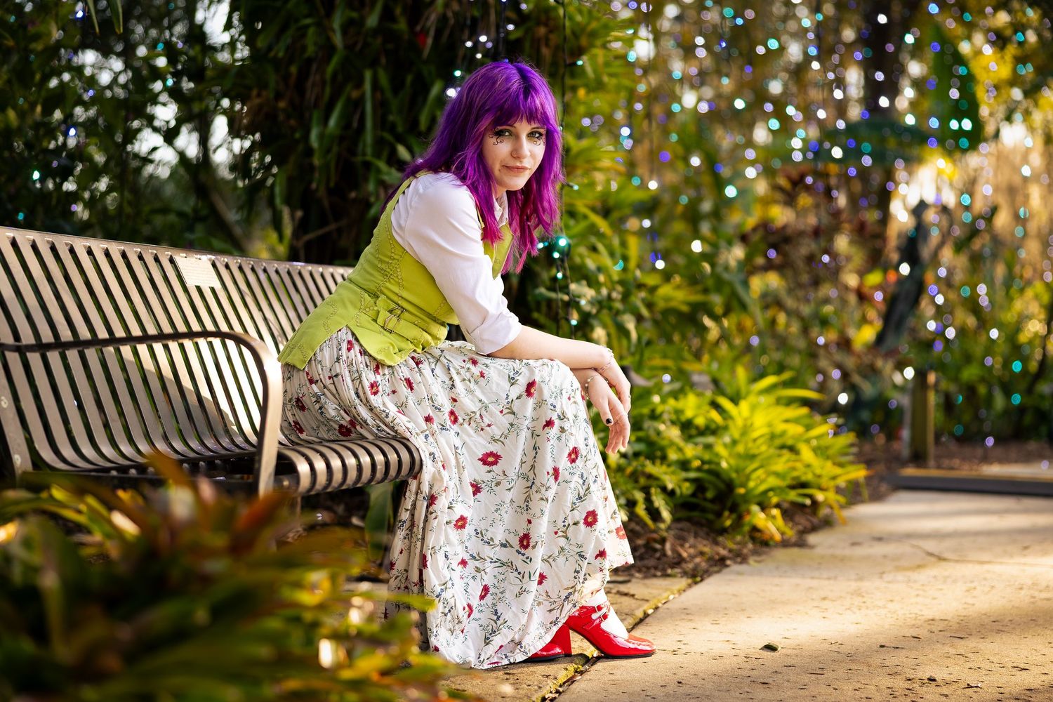 Woman with purple hair in floral skirt and green vest sits on park bench surrounded by lush greenery.