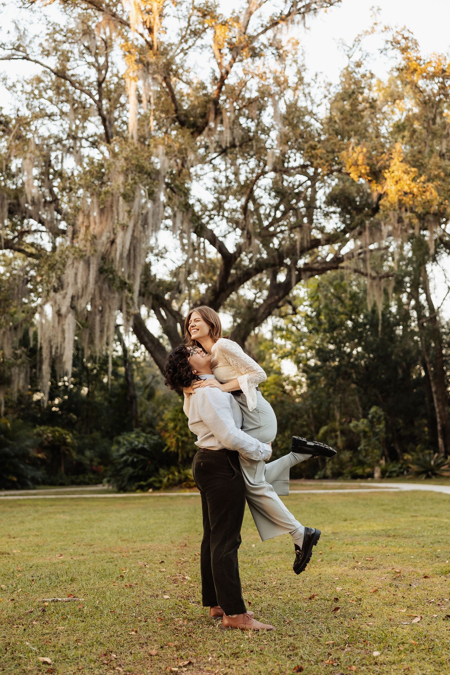 One woman joyfully lifts her partner in a sunny park beneath Spanish moss-draped oak trees.