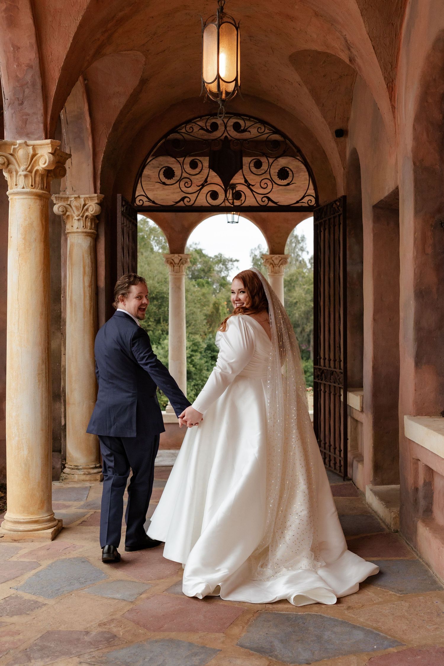 Bride and groom hold hands under ornate arched colonnade of a historic Spanish venue.