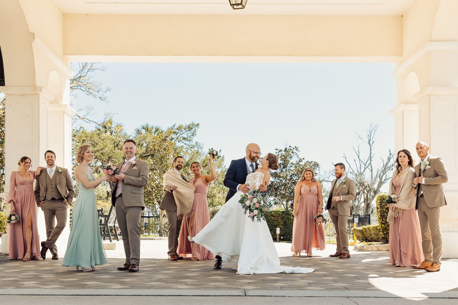 Groom dipping bride surrounded by wedding party in mauve and sage dresses under a portico.