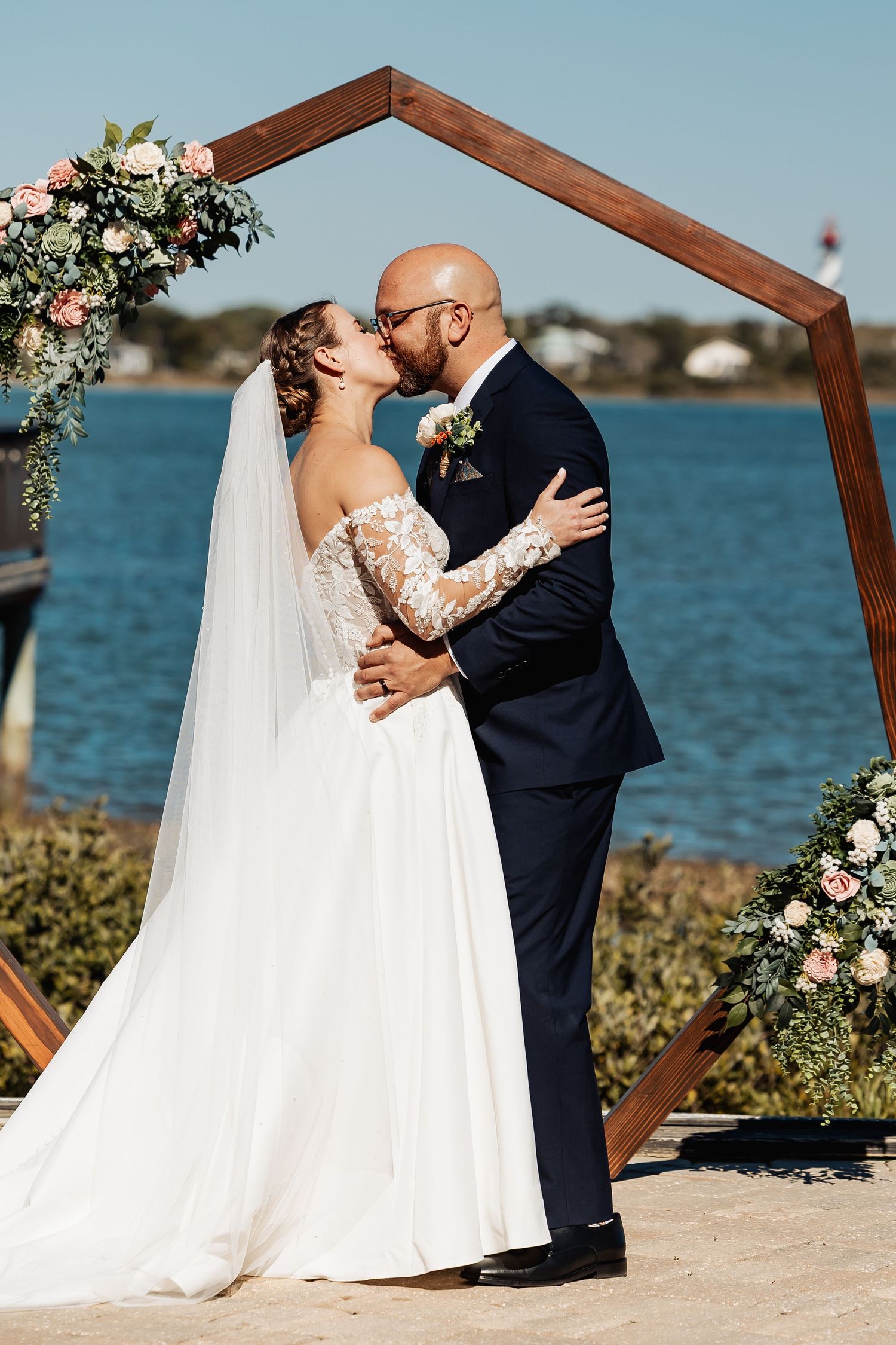 Bride and groom share first kiss under wooden hexagonal arch adorned with flowers by the waterfront.