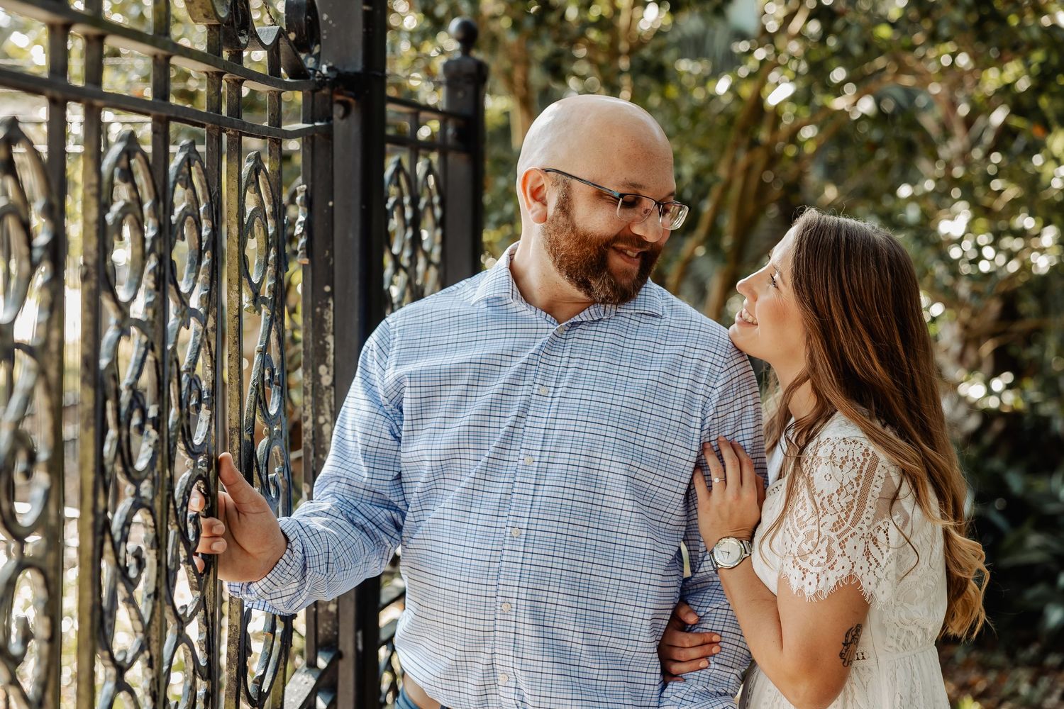 Couple smiling at each other while standing near ornate black iron gate outdoors.
