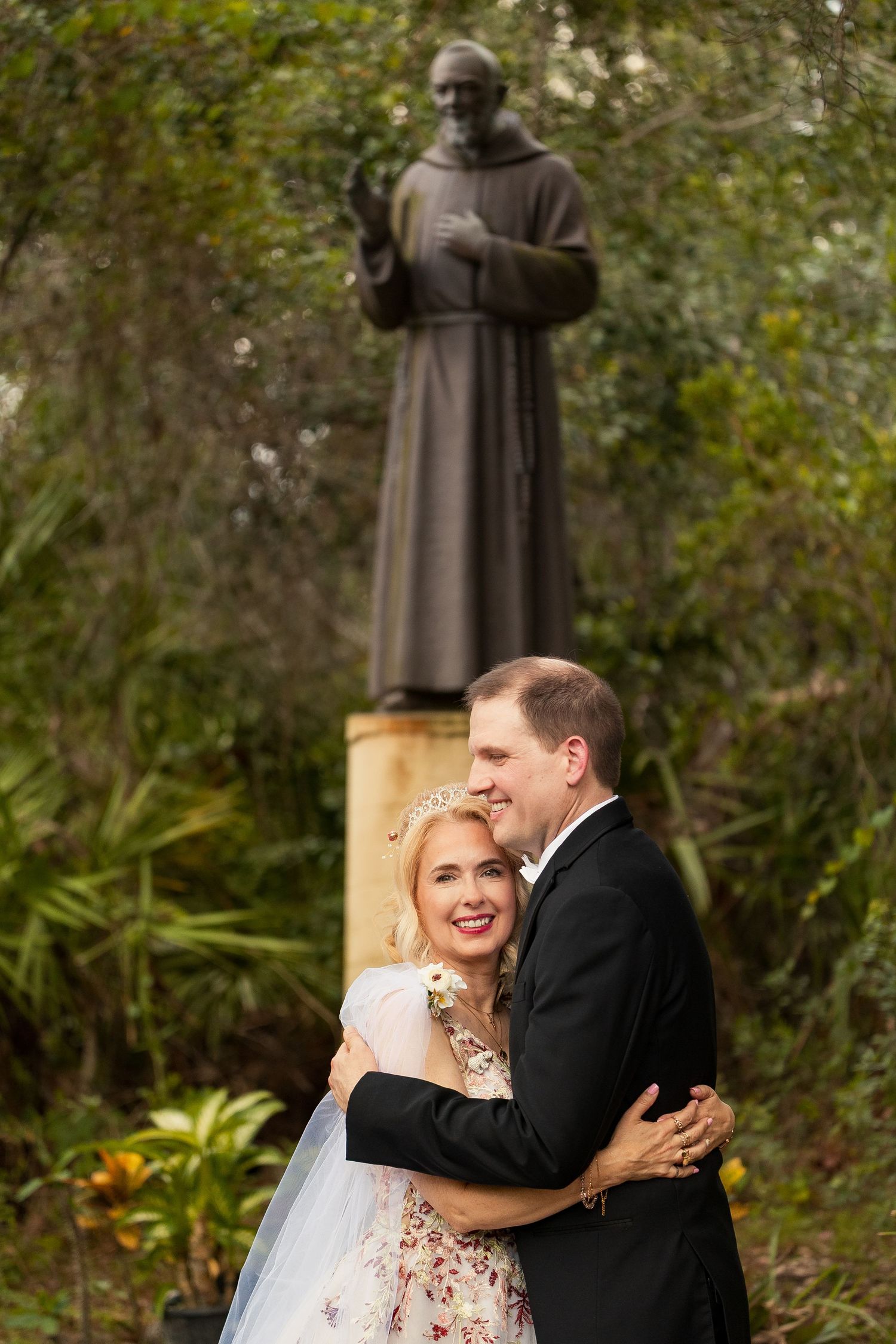Happy couple embracing in front of a Saint Francis statue in a lush garden wedding setting.