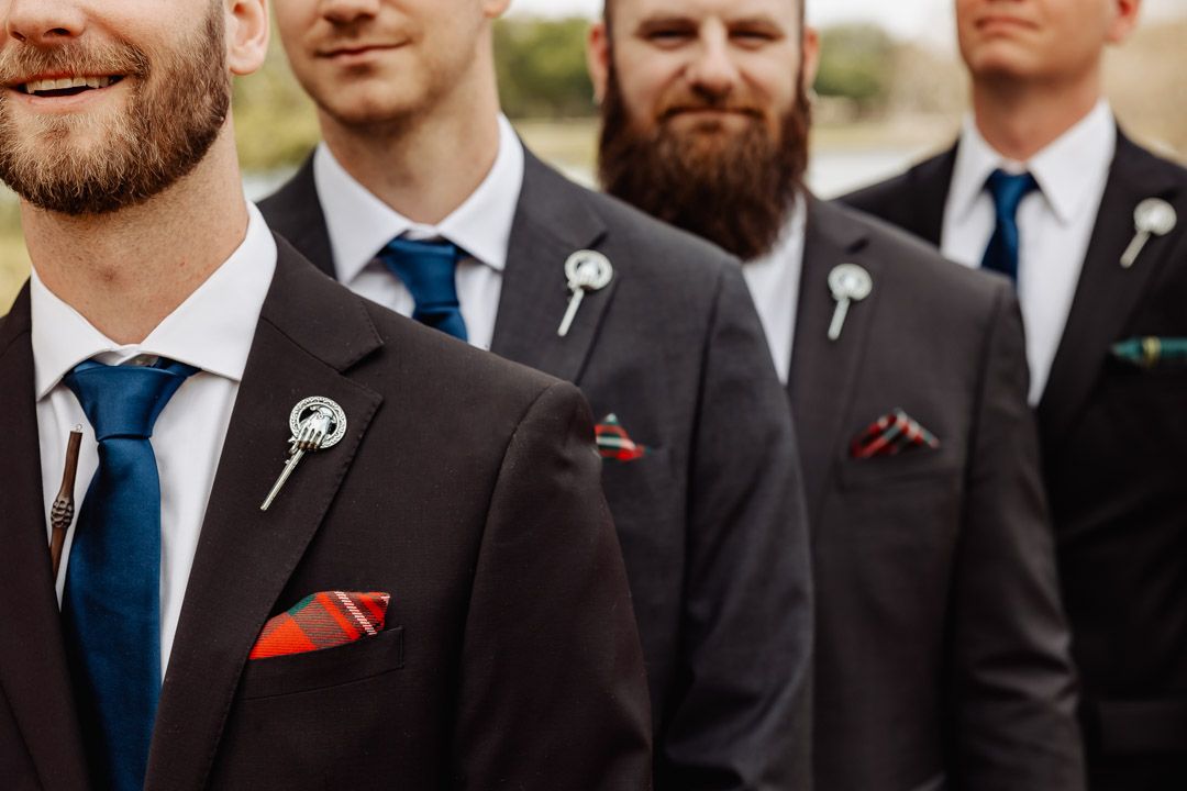 Close-up of groomsmen in dark suits with blue ties and Scottish clan badge boutonnieres at wedding.