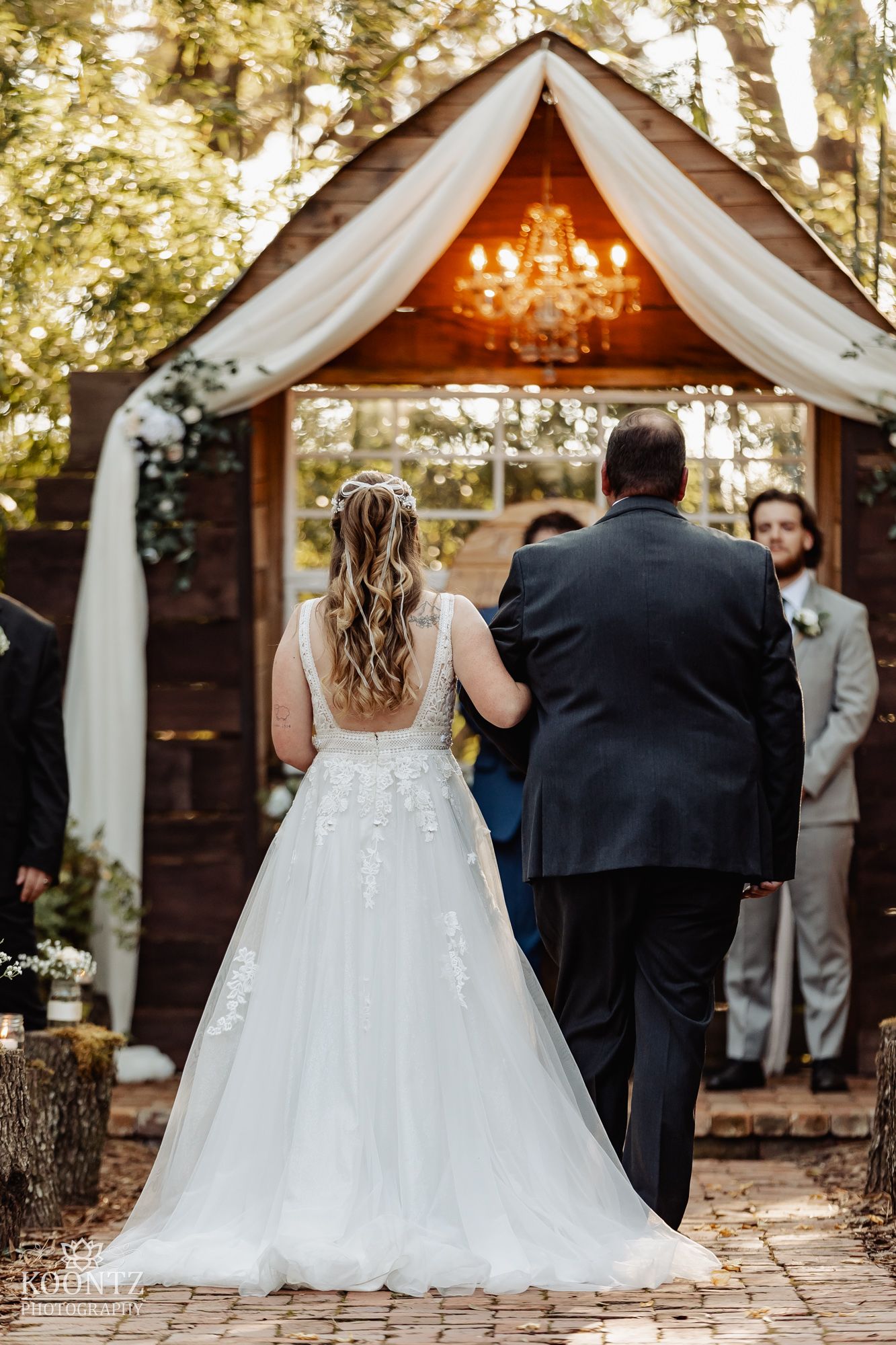 Bride in floral lace wedding gown walks down brick aisle toward rustic wooden altar with chandelier.