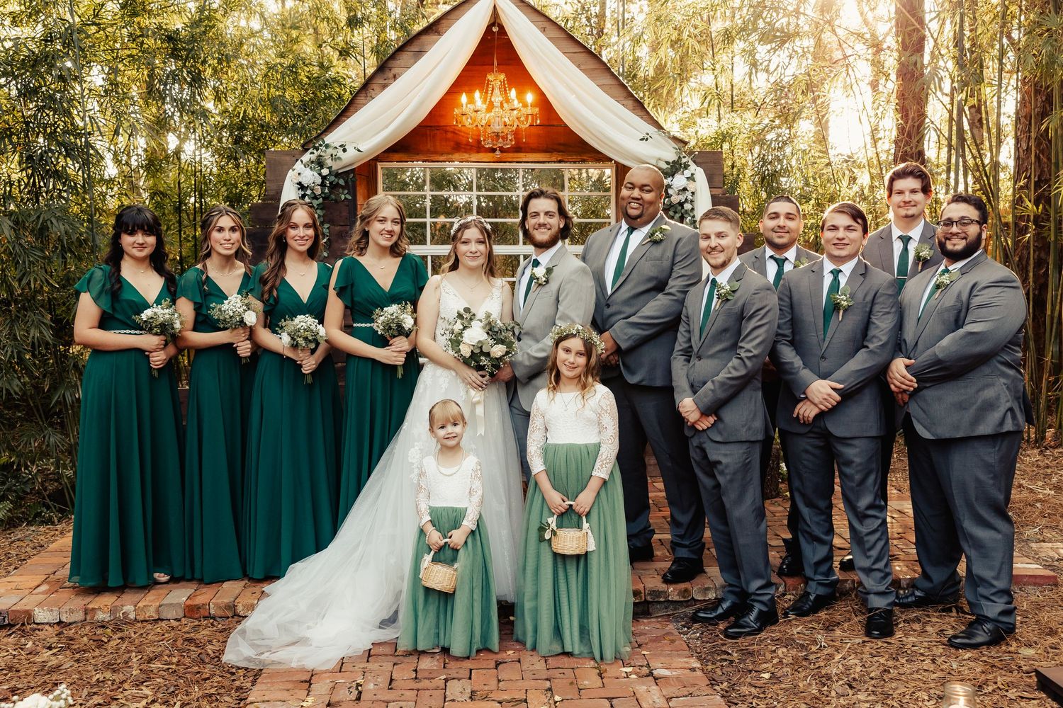 Wedding party in emerald green and grey poses in front of wooden chapel in forest setting.