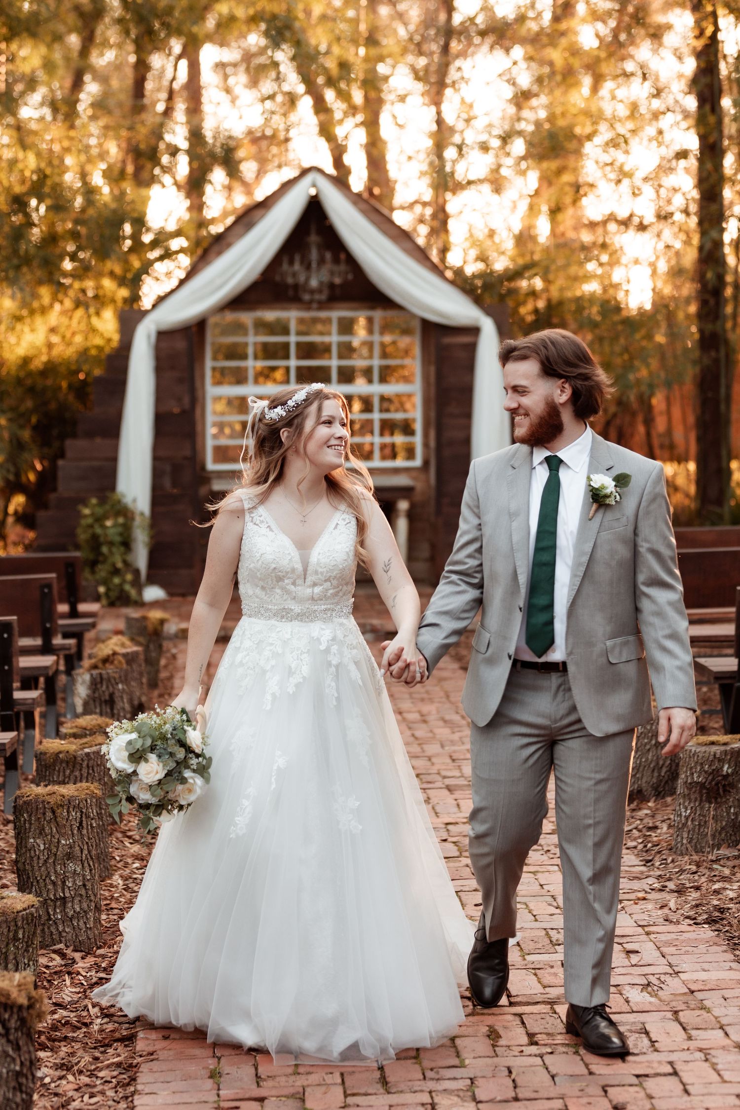 Newlyweds walk hand in hand on brick path with rustic chapel and autumn foliage in background.