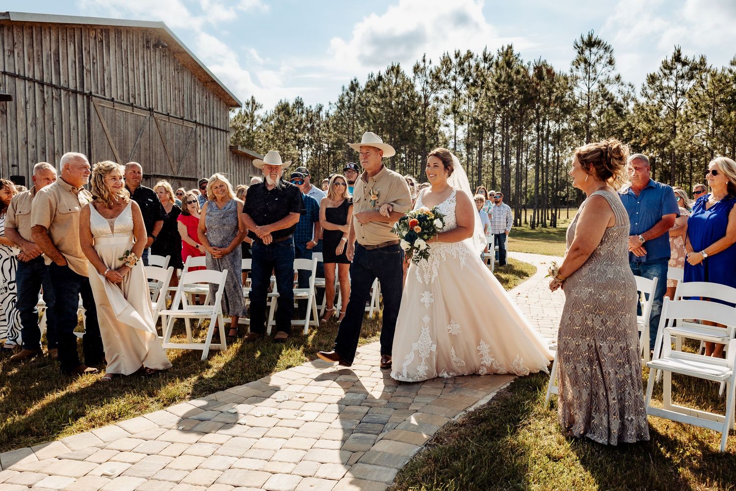 Wedding ceremony guests gathered outside rustic barn on stone pathway.