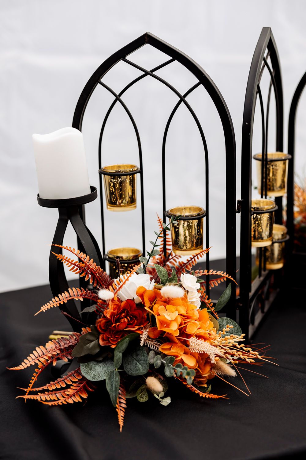 Gothic black arched candle holder with orange fall floral arrangement and gold votives on black tablecloth.
