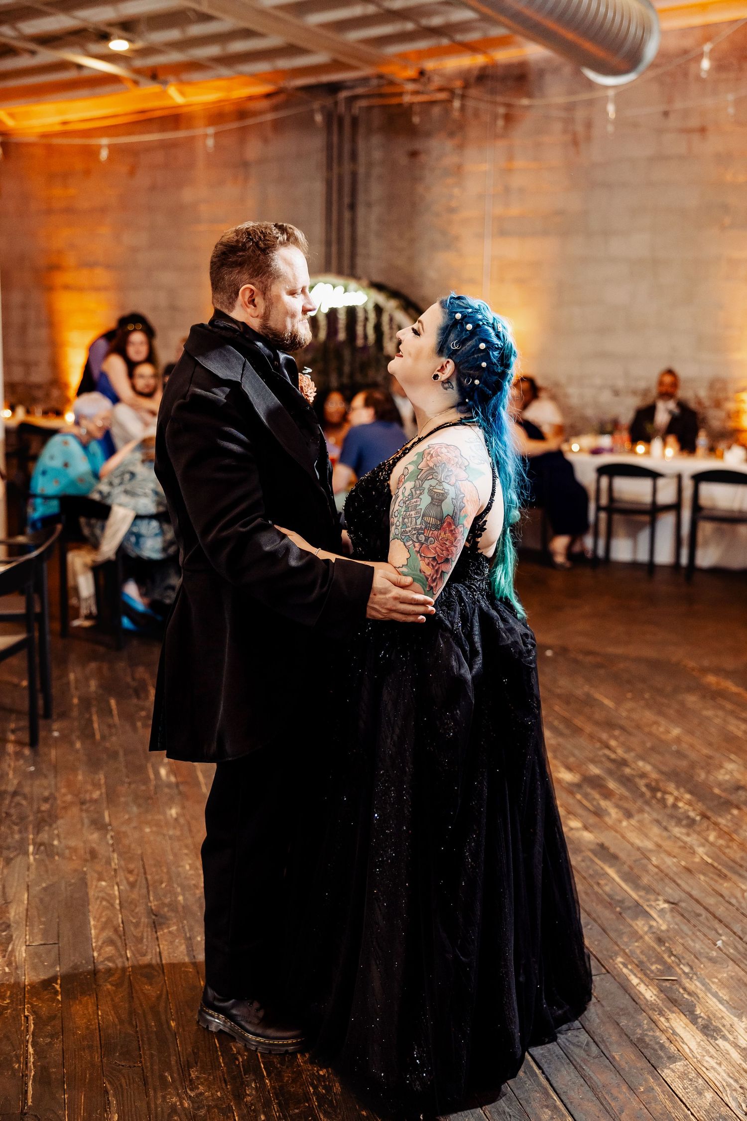 Tattooed bride in black gown and groom in black velvet suit share first dance at wedding reception.