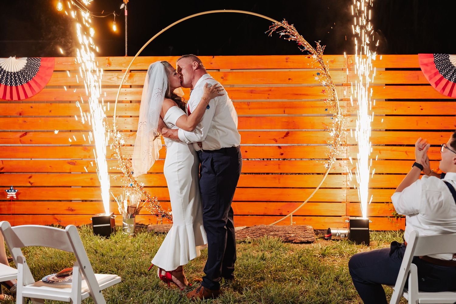 Bride and groom share first kiss with sparkler fountains exploding beside circular arch at outdoor night wedding.