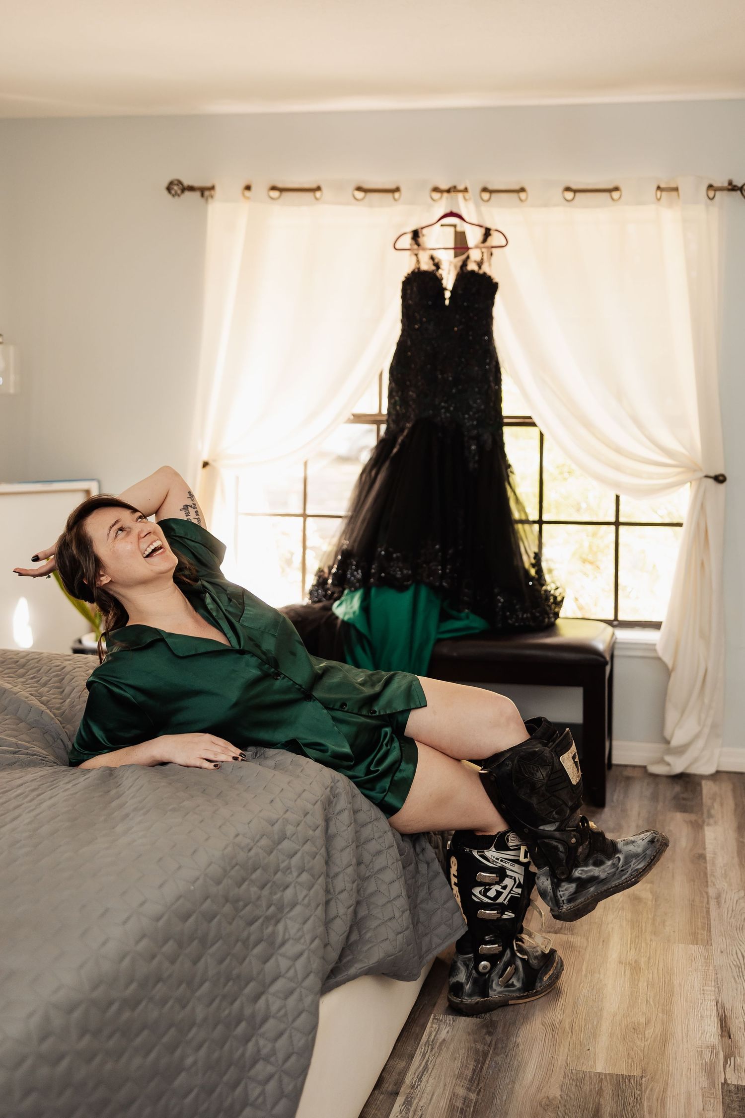 A woman in a green robe laughing on a bed with a black wedding gown hanging behind her.