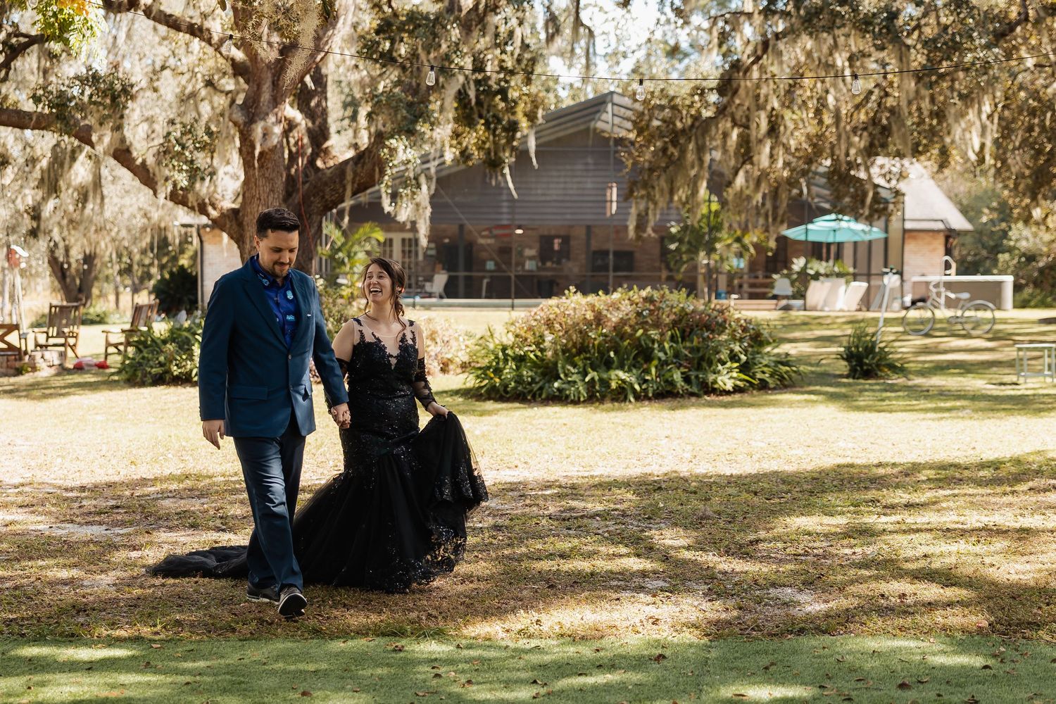 Couple walks through moss-draped oak trees, bride in black lace gown and groom in navy suit at outdoor wedding.