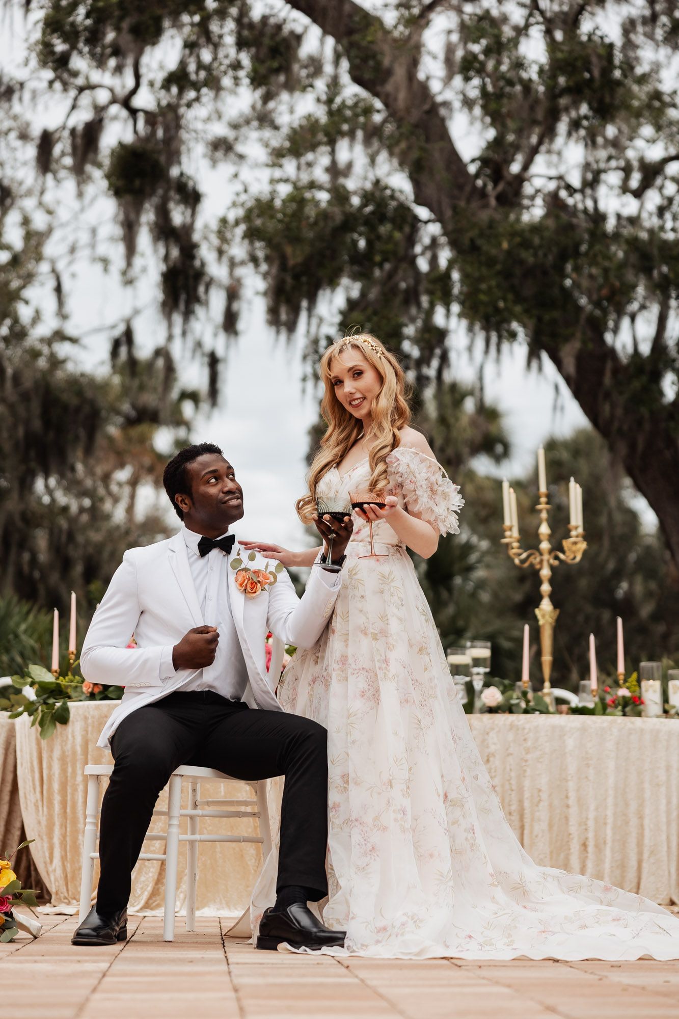 Groom in white tuxedo and bride in lace gown toasting wine glasses at outdoor wedding reception.