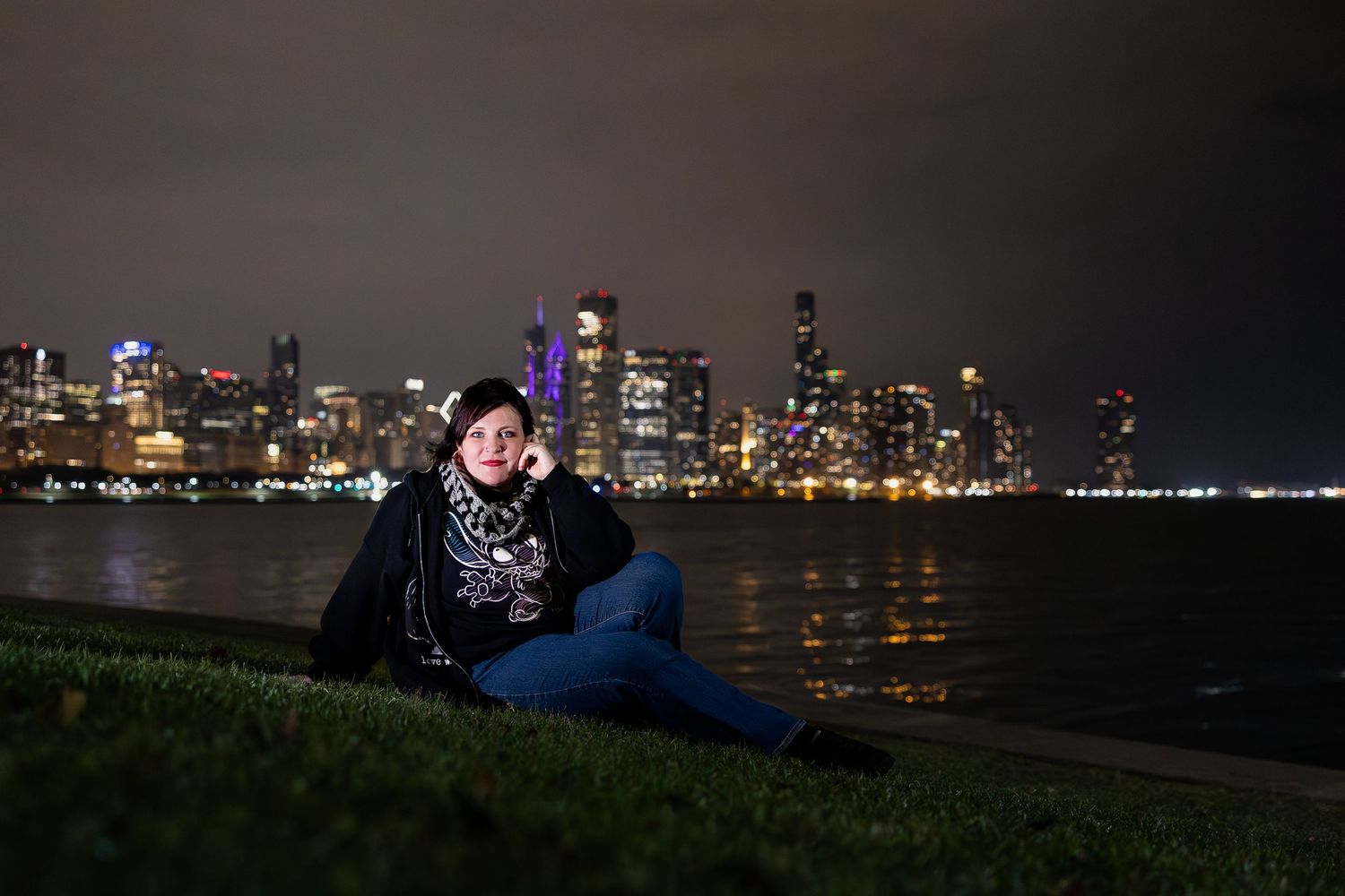 Person sitting on grass at night with illuminated city skyline reflecting on water in background.