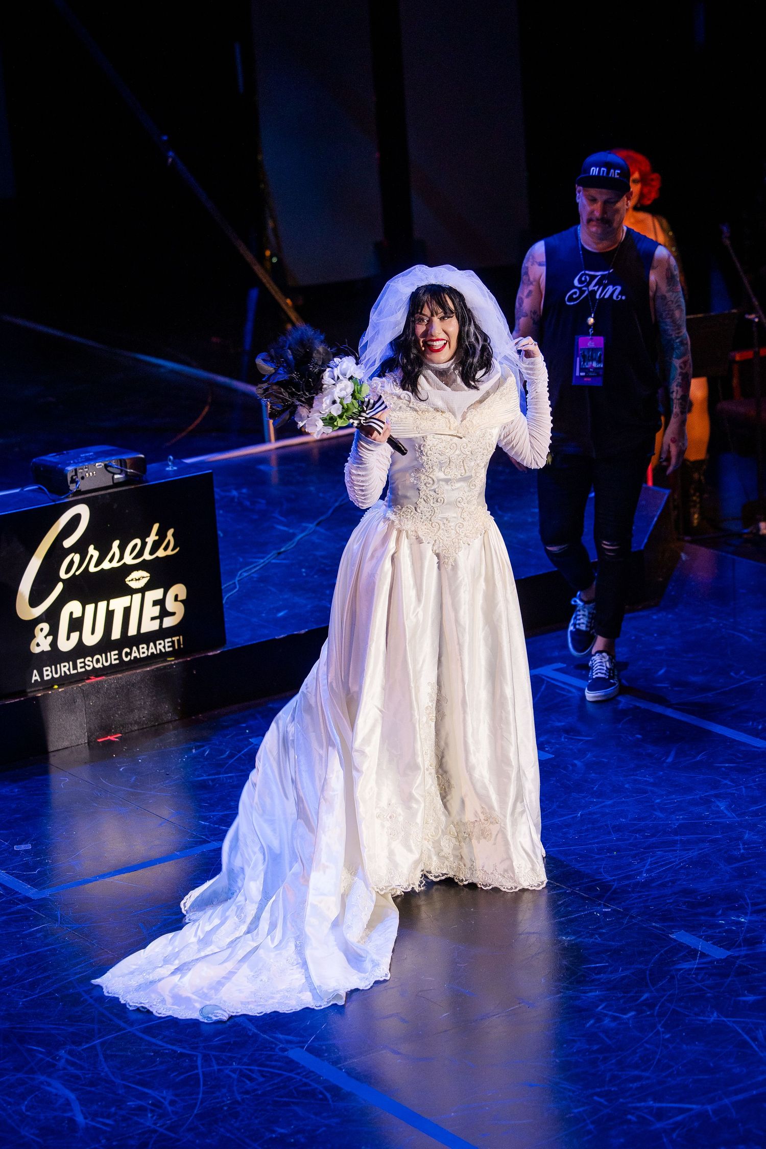 A performer in a white wedding gown and veil holds a bouquet on the Corsets and Cuties Cabaret stage.