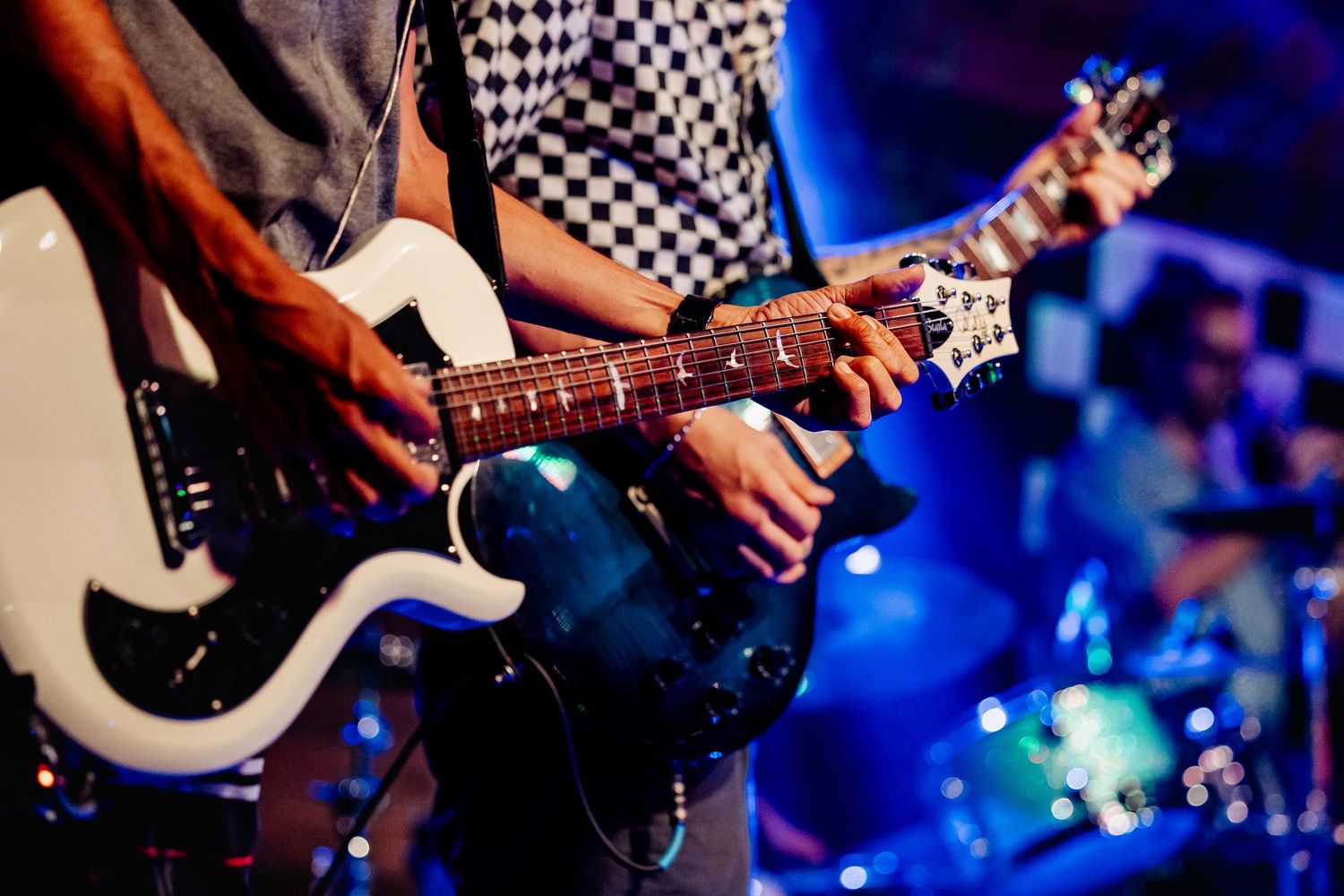 Close-up of two electric guitarists playing on stage under blue concert lighting with drummer behind.