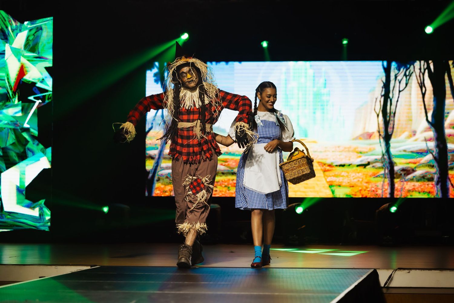 Dorothy and Scarecrow Wizard of Oz costumed models walk runway with scenic backdrop display.