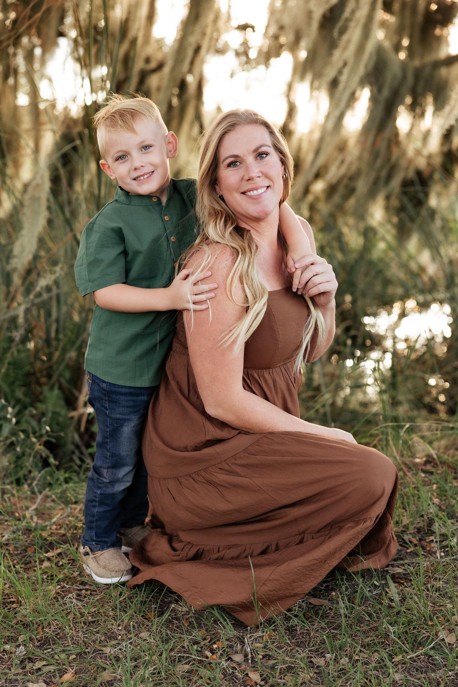 Smiling blonde mother kneeling with young son hugging her from behind under Spanish moss trees.