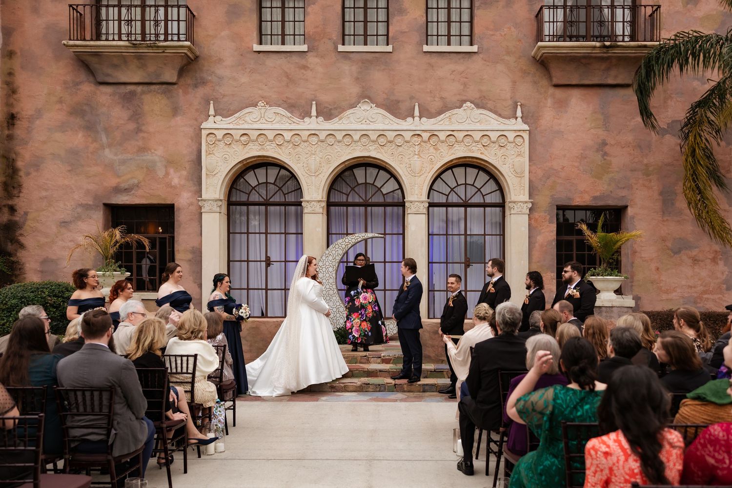 Outdoor wedding ceremony at elegant Spanish-style venue with bride and groom at floral altar.