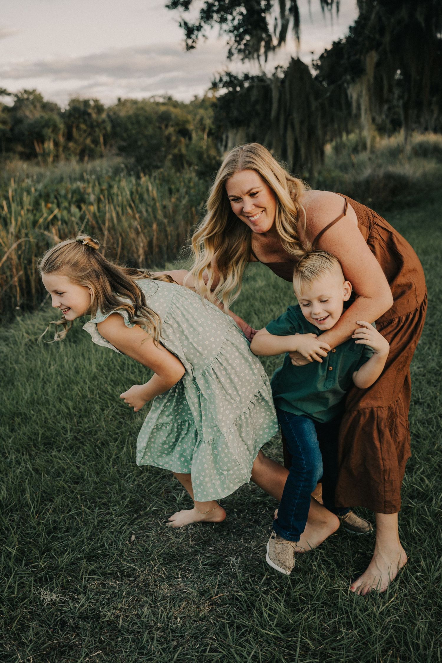 Playful outdoor scene showing a barefoot family in brown and green outfits running together on grass.