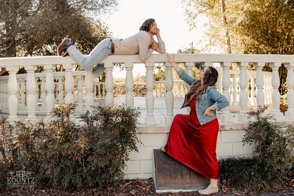 Playful couple posing by white balustrade with woman in red dress and man lying on railing.