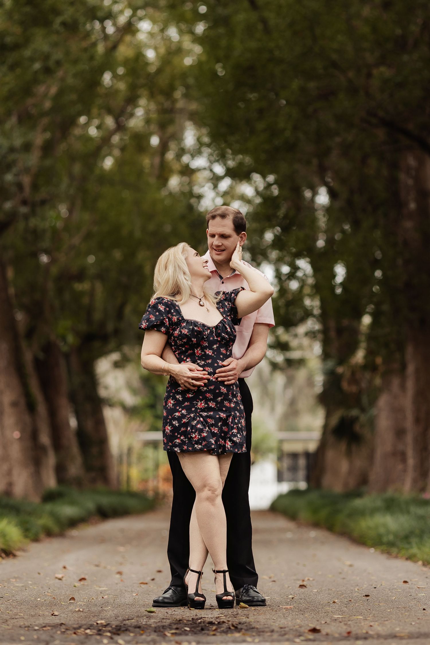 Couple embracing and smiling at each other on a tree-lined pathway in a botanical garden.