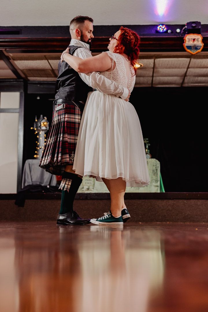 Bride in white tea-length dress and Converse slow dancing with groom in tartan kilt at reception.