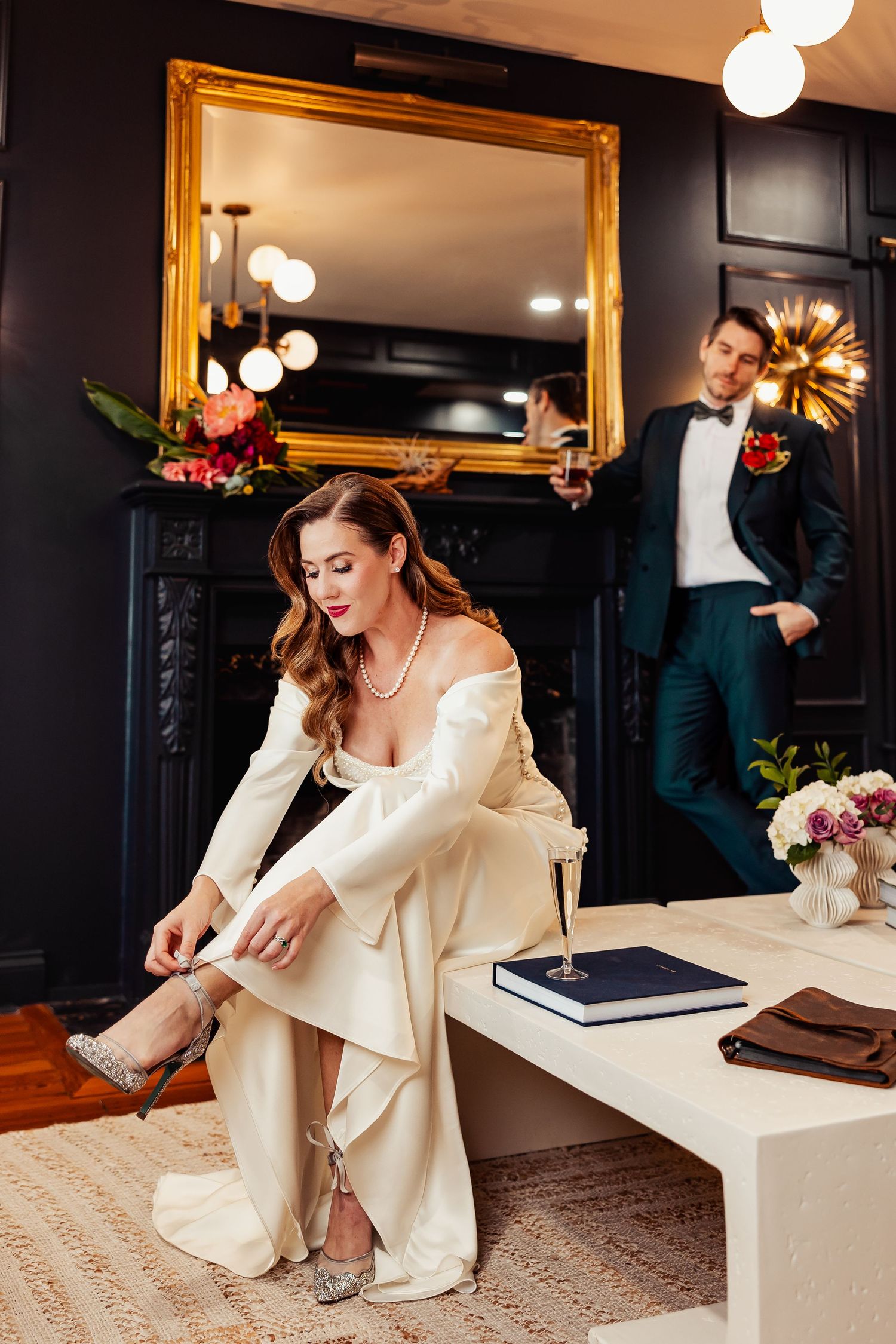 Bride putting on heels in elegant dark room as groom stands by ornate gold mirror.