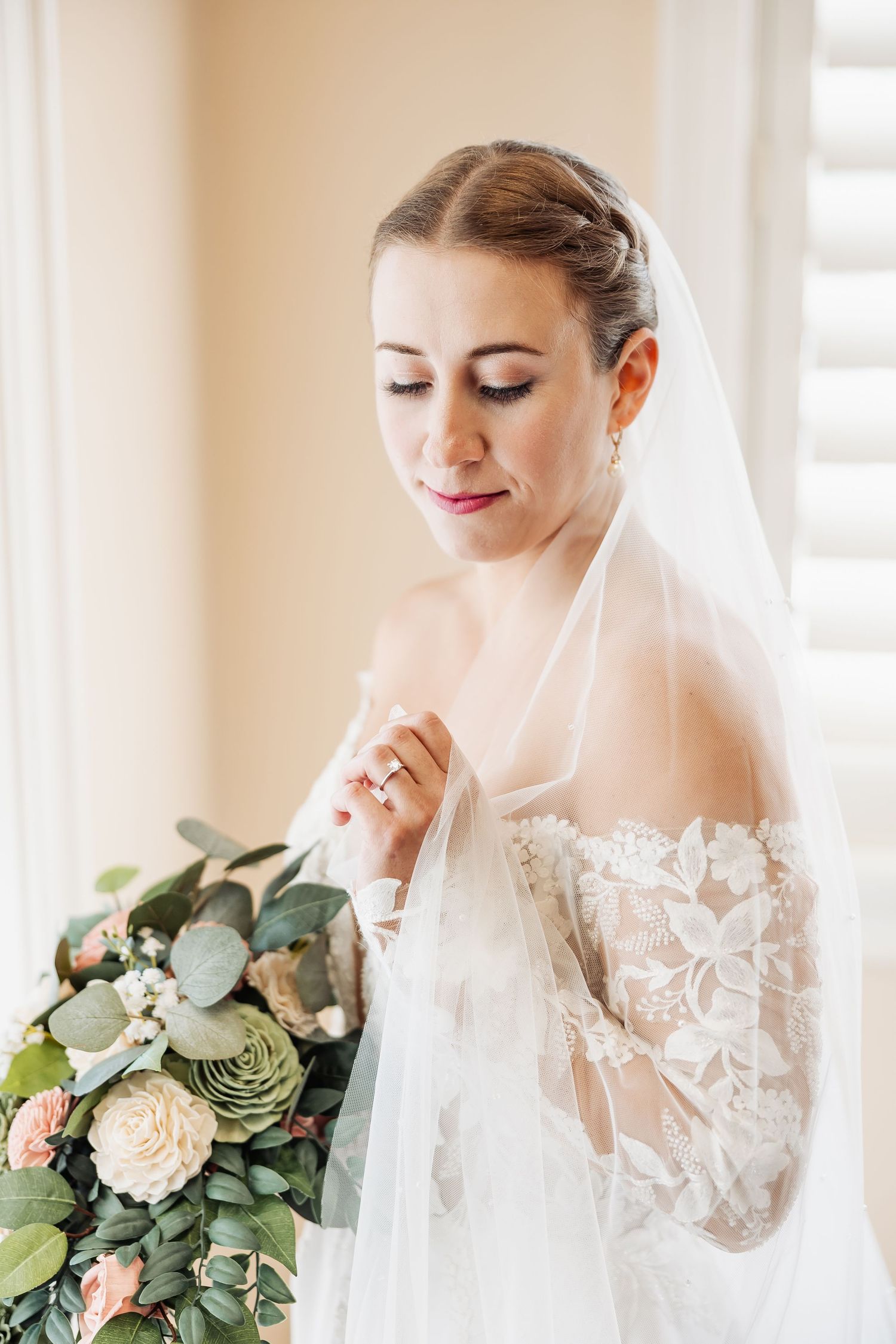 Elegant bride in lace gown with veil holding a bouquet of pink and green flowers.