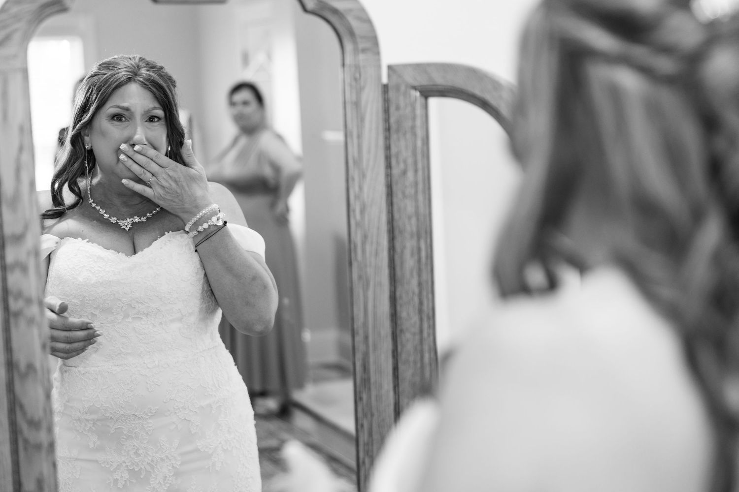 Emotional bride in white lace gown covering her mouth while looking at her reflection in a mirror.