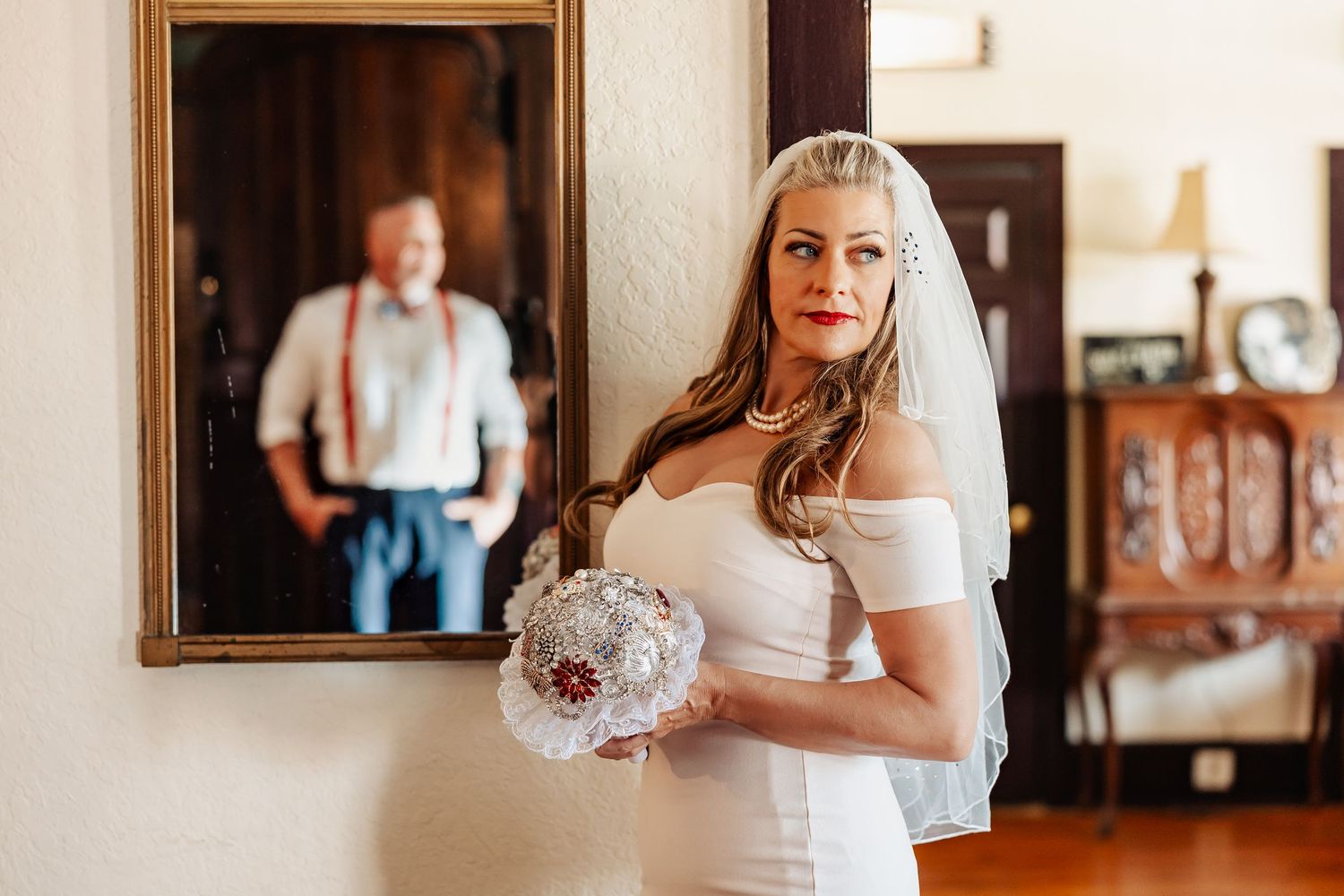 Bride in white dress holding bouquet with groom reflected in mirror behind her.