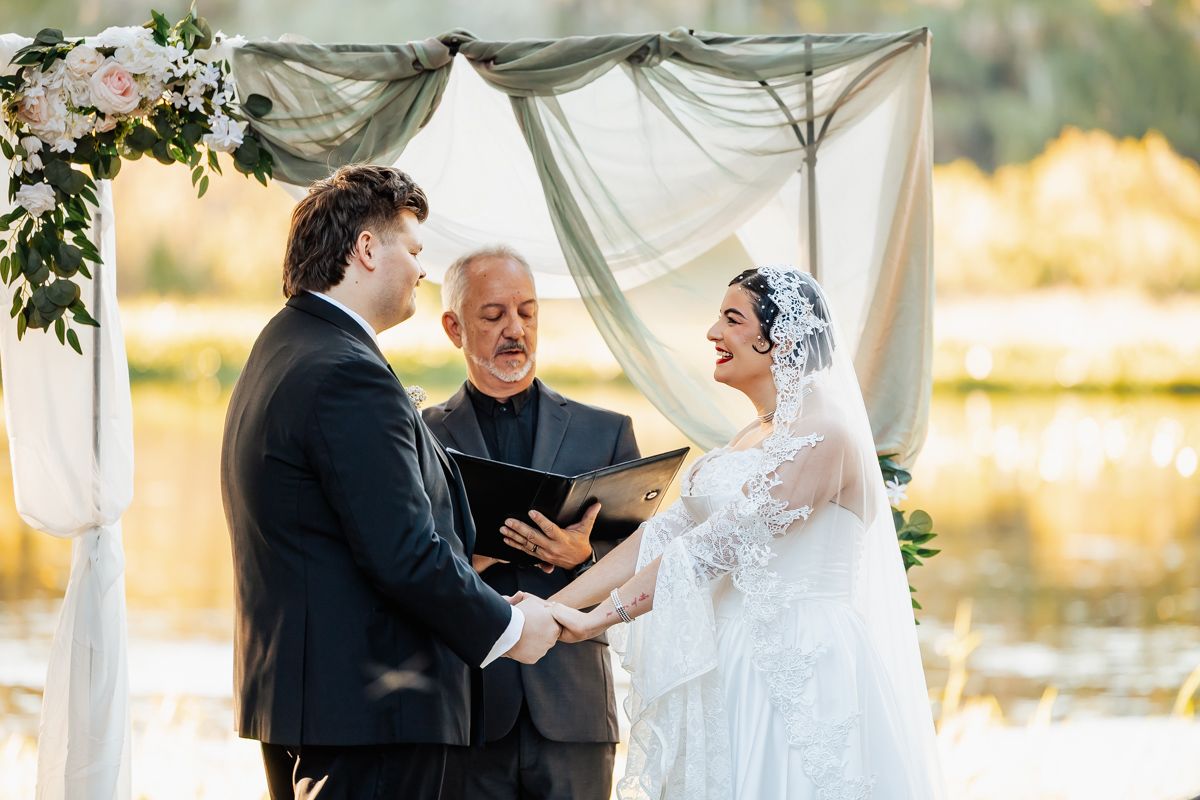 Bride and groom exchange vows under draped altar with officiant by scenic lakeside at sunset.