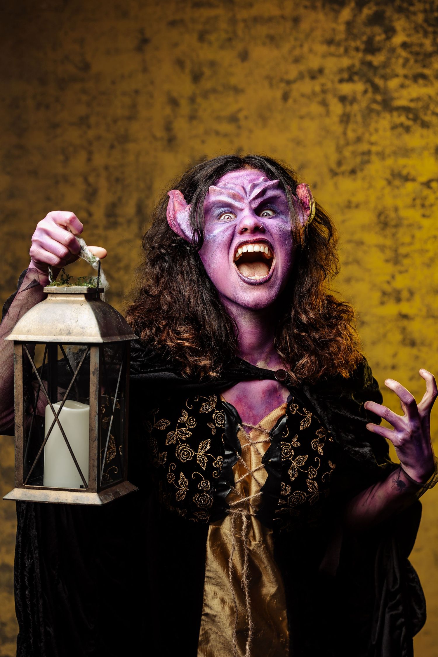 Dramatic creature makeup with pointed ears and fangs, holding lantern against yellow backdrop.