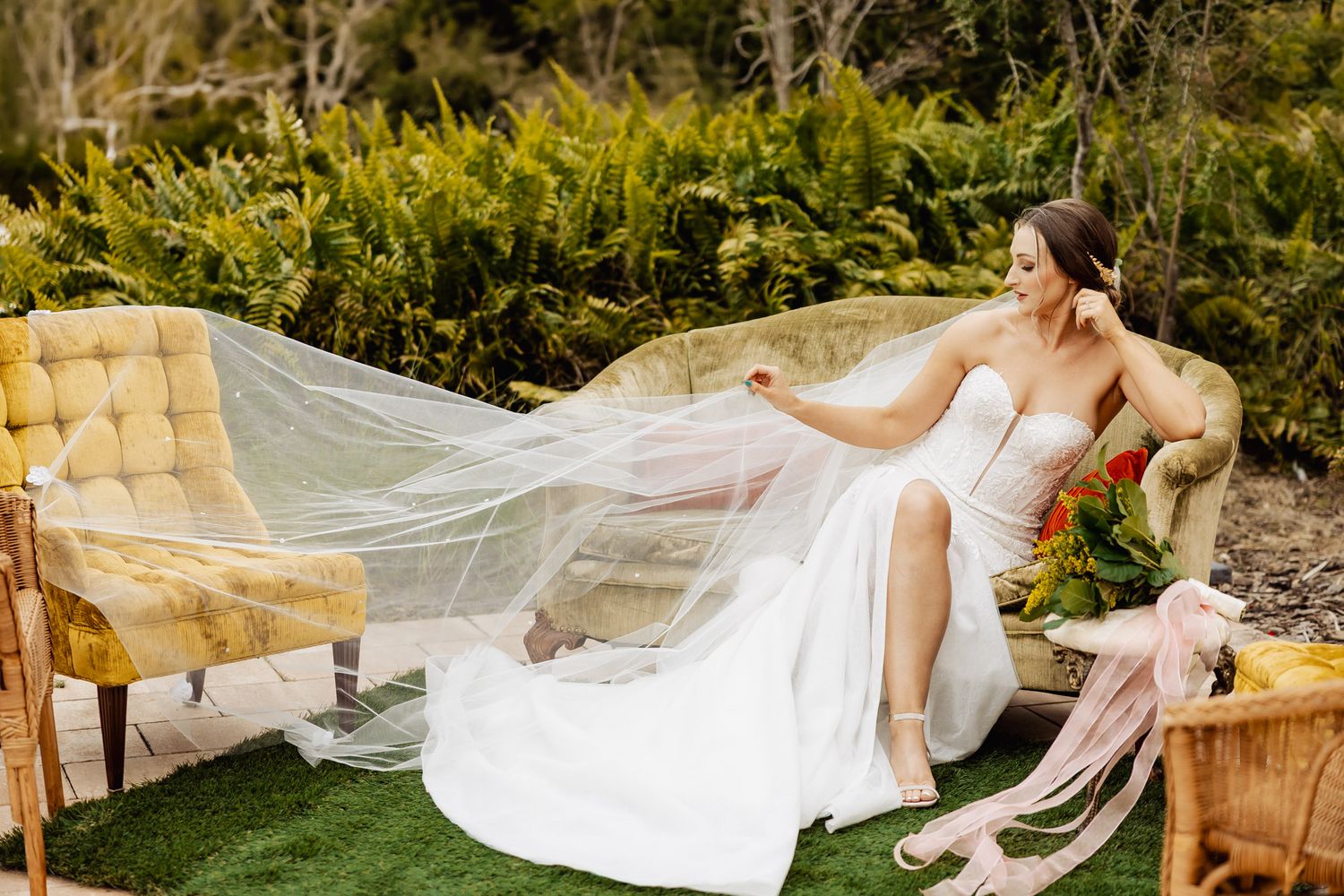 Bride in white gown and flowing veil reclines on vintage yellow sofa outdoors surrounded by lush greenery.
