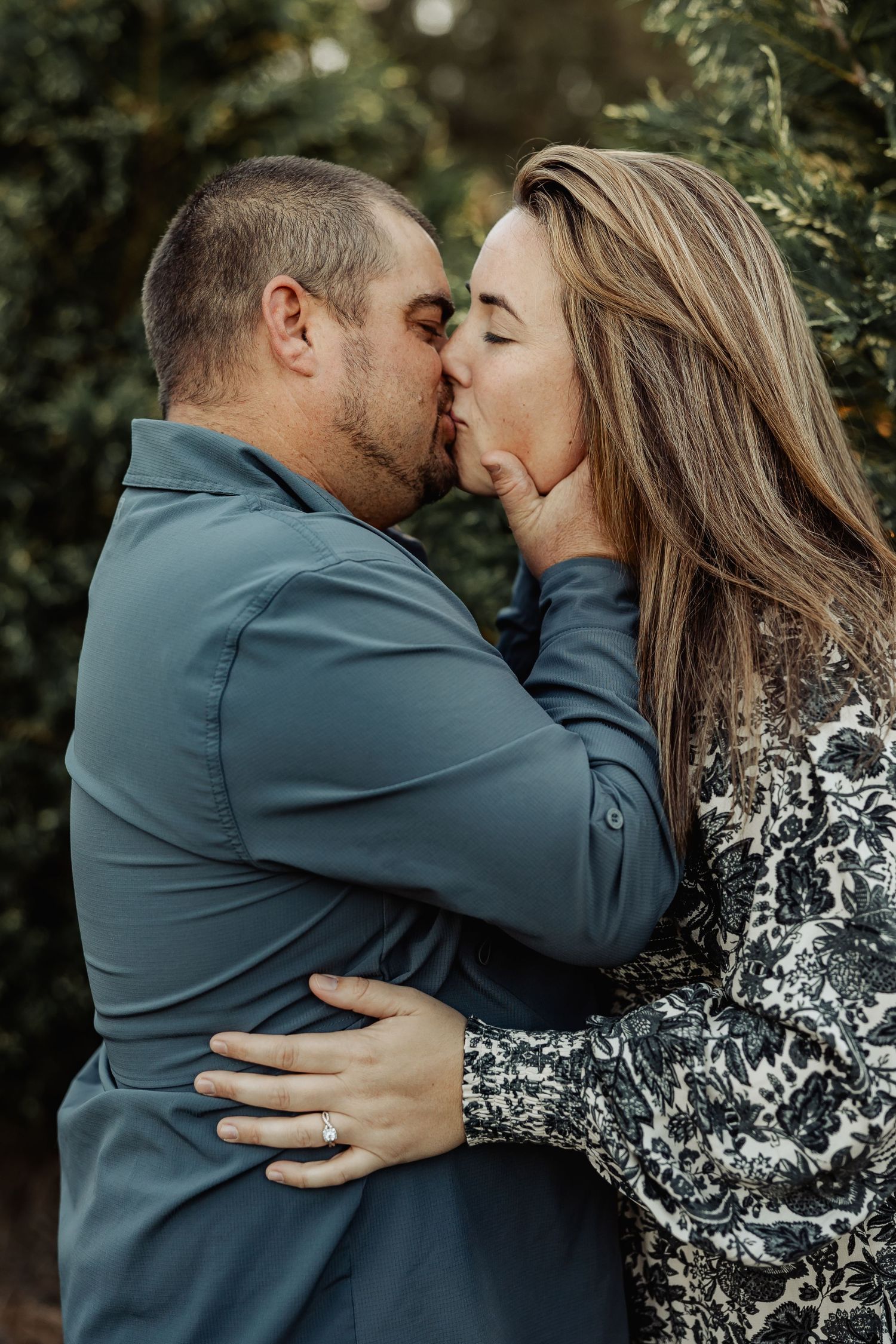 An engaged couple shares a passionate kiss outdoors, with the woman's diamond ring visible.