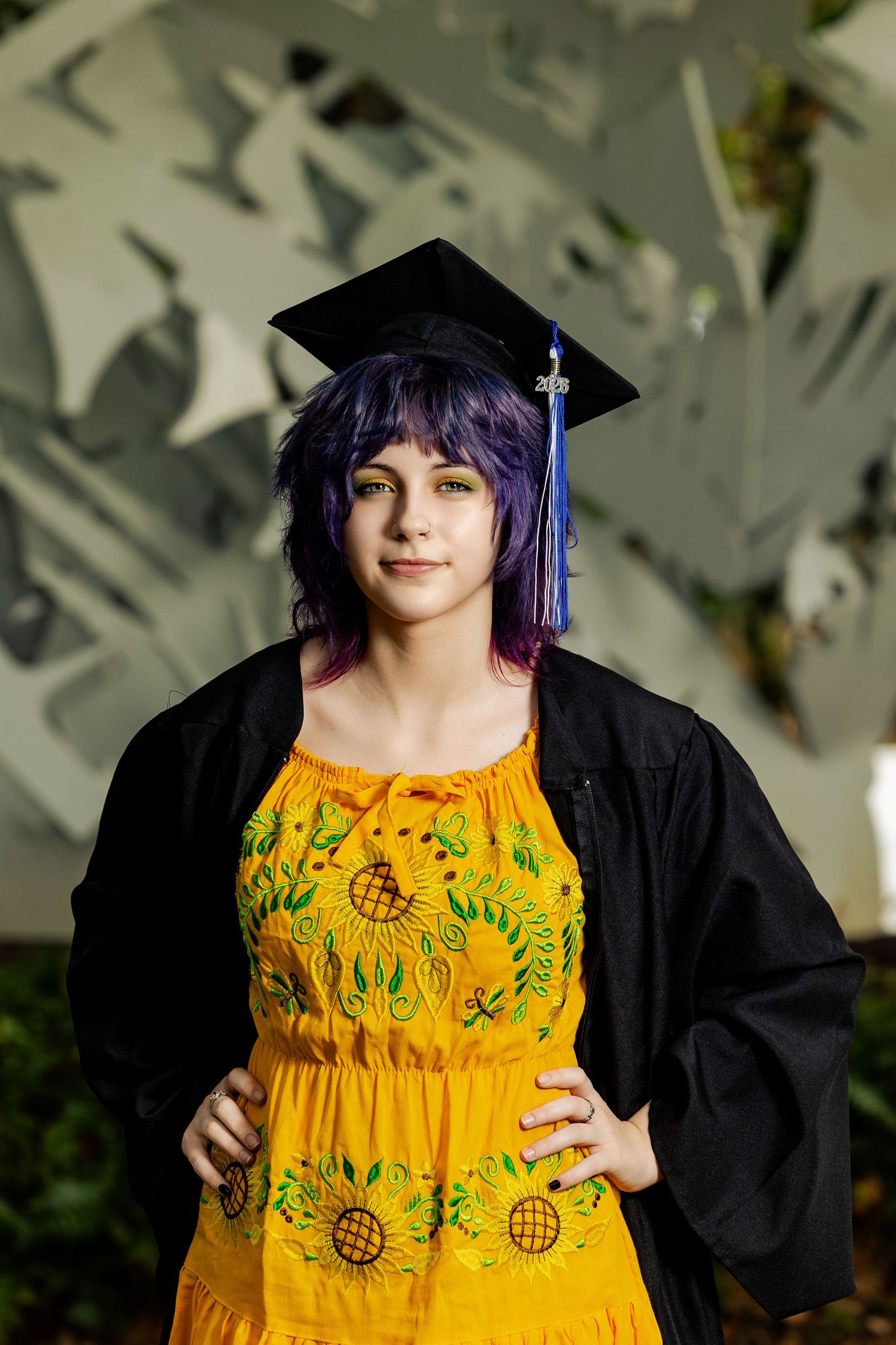 Young woman with purple hair wearing a yellow sunflower dress and black graduation cap and gown poses confidently.