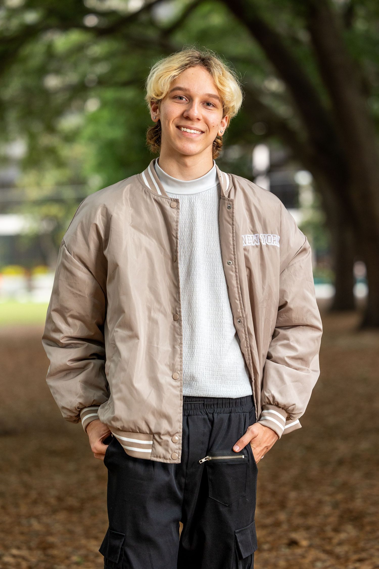 Young man with blonde hair smiling outdoors, wearing a tan bomber jacket and black cargo pants in a tree-lined park.
