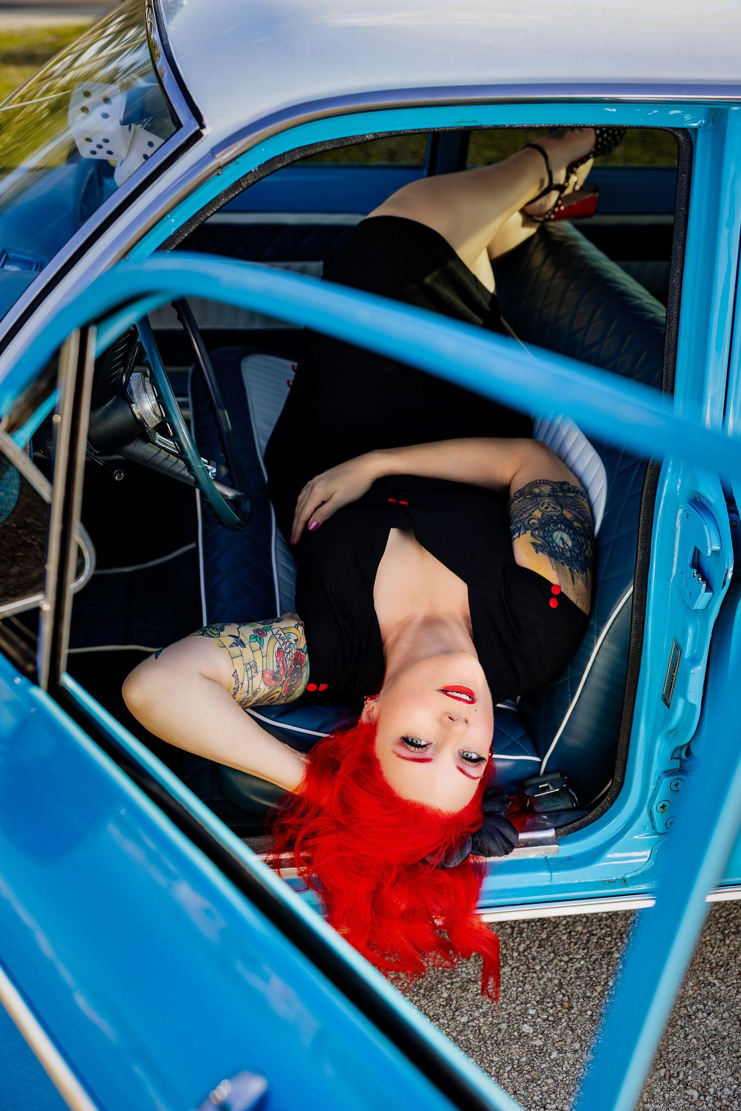 Red-haired tattooed woman in black outfit posing reclined inside a vintage blue classic car with dice hanging from mirror.