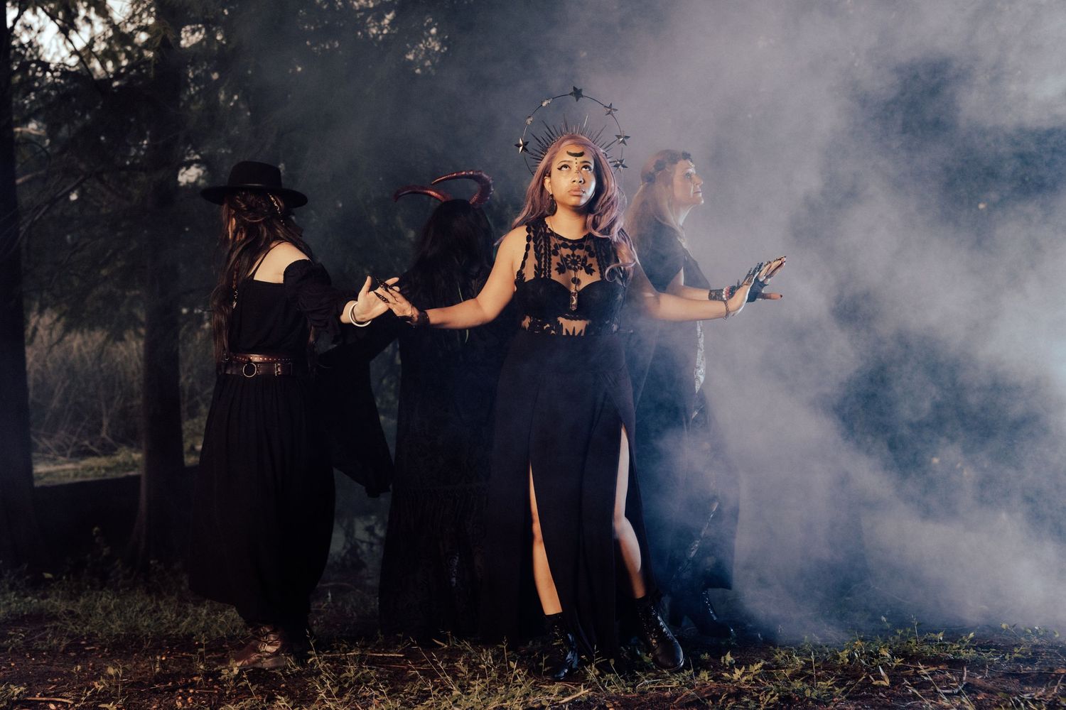 Four women in black witch costumes pose dramatically in a misty forest during a Halloween photoshoot.