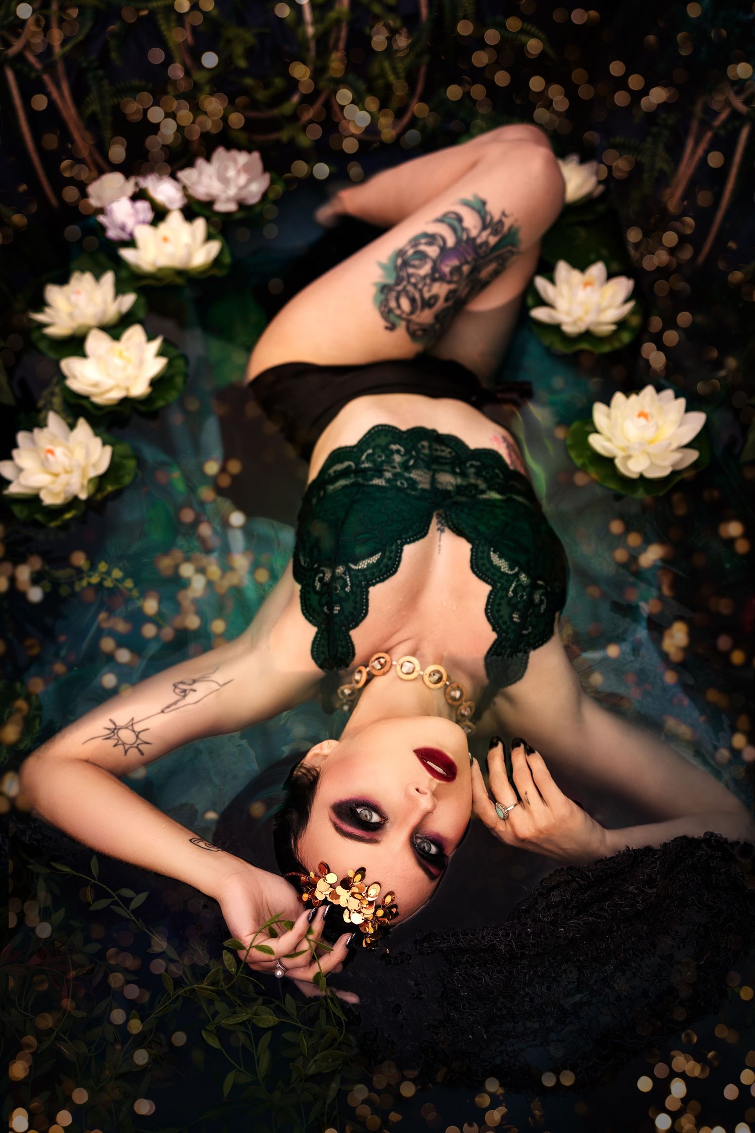 Tattooed woman in green lace lingerie lies among white water lilies holding gold flower accessory.
