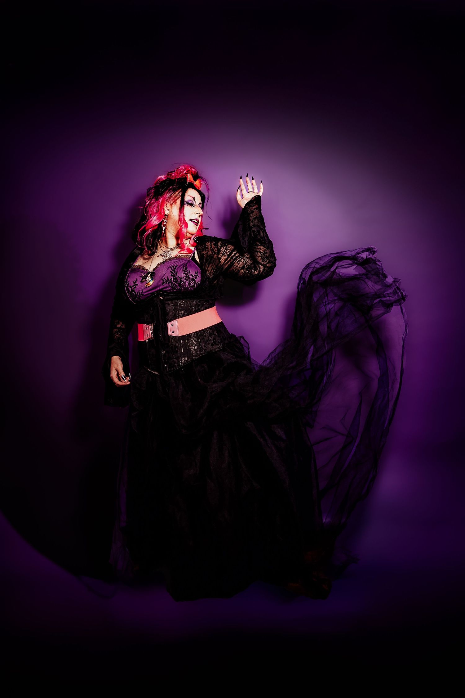 Woman in gothic Halloween costume with pink hair, black lace dress, and dramatic makeup poses against purple background.