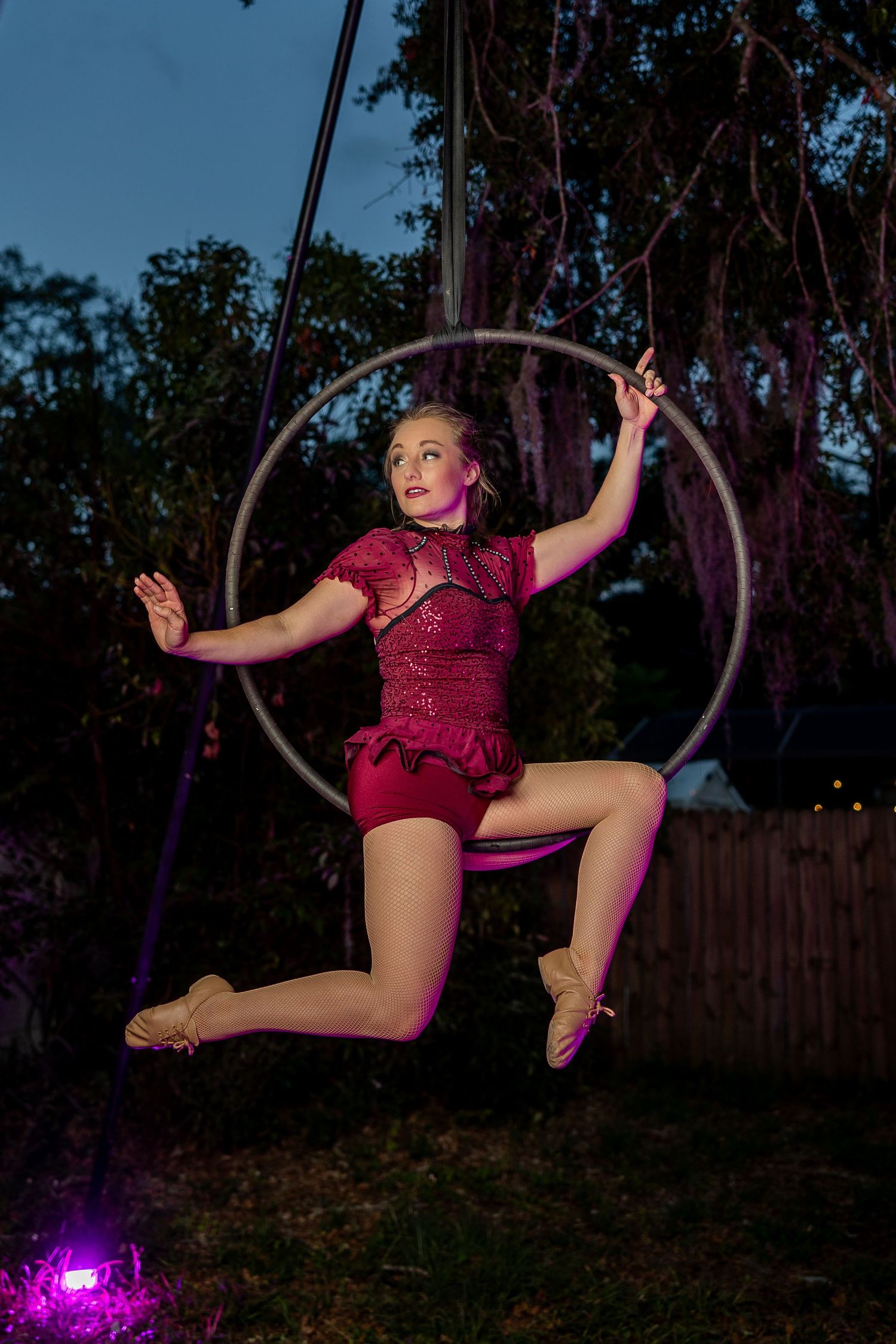 Young aerialist in red costume performs on a lyra hoop at dusk with purple lighting.