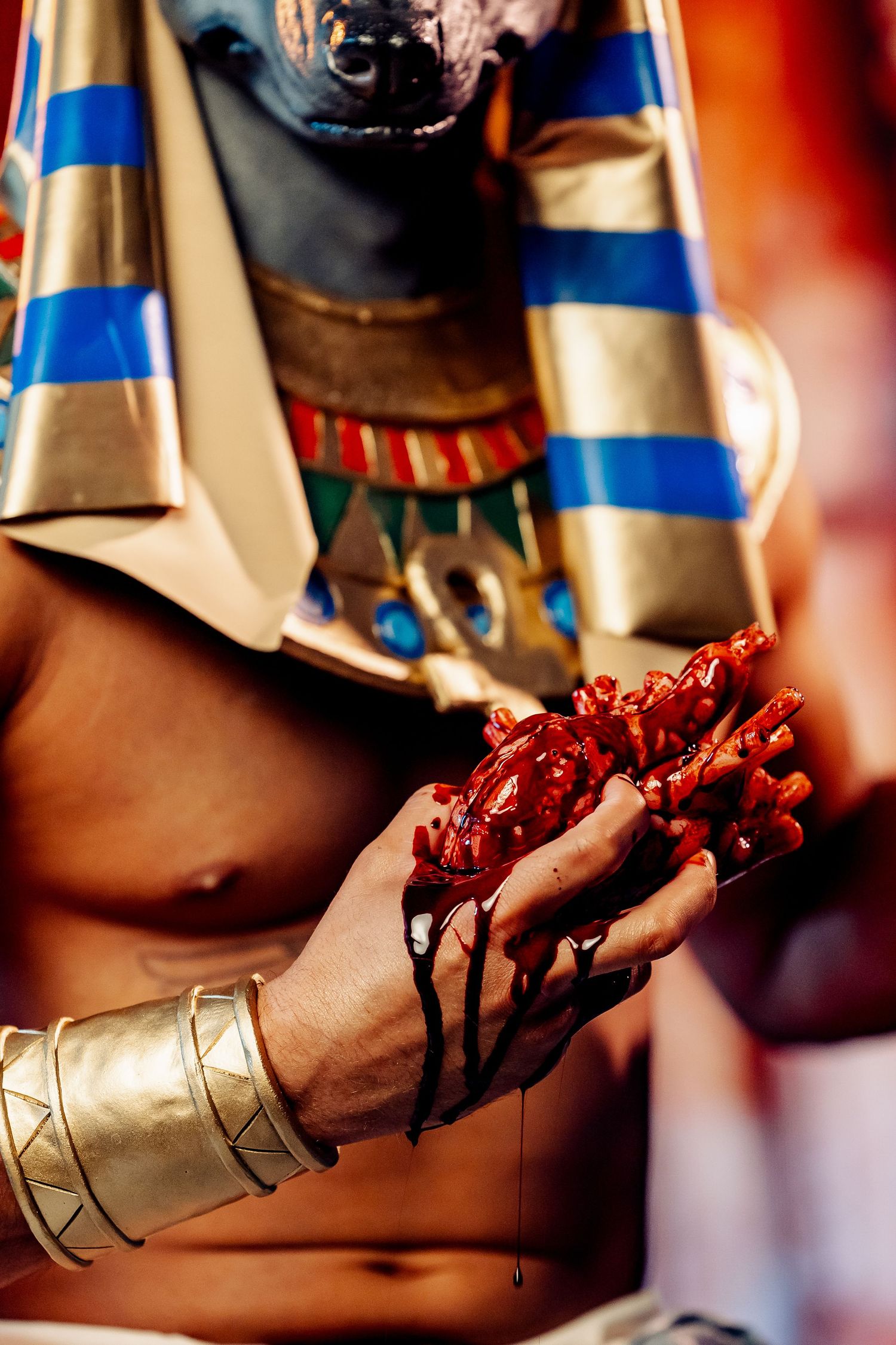 Person in Egyptian Anubis costume holding bloody prop organs with gold bracelet and striped headdress.
