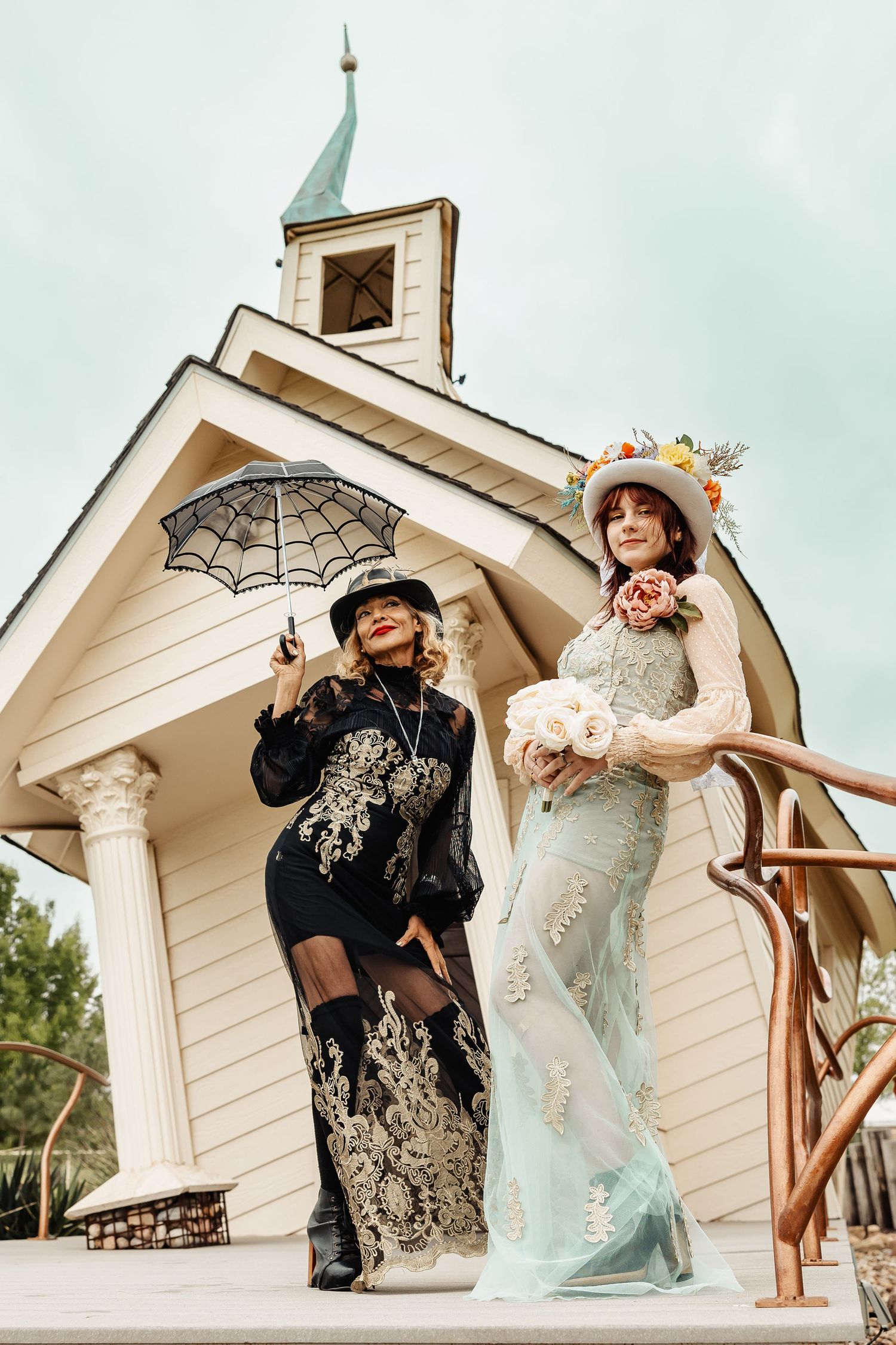 Two elegantly dressed women pose outside white chapel, one holding spider web umbrella, other carrying floral bouquet.