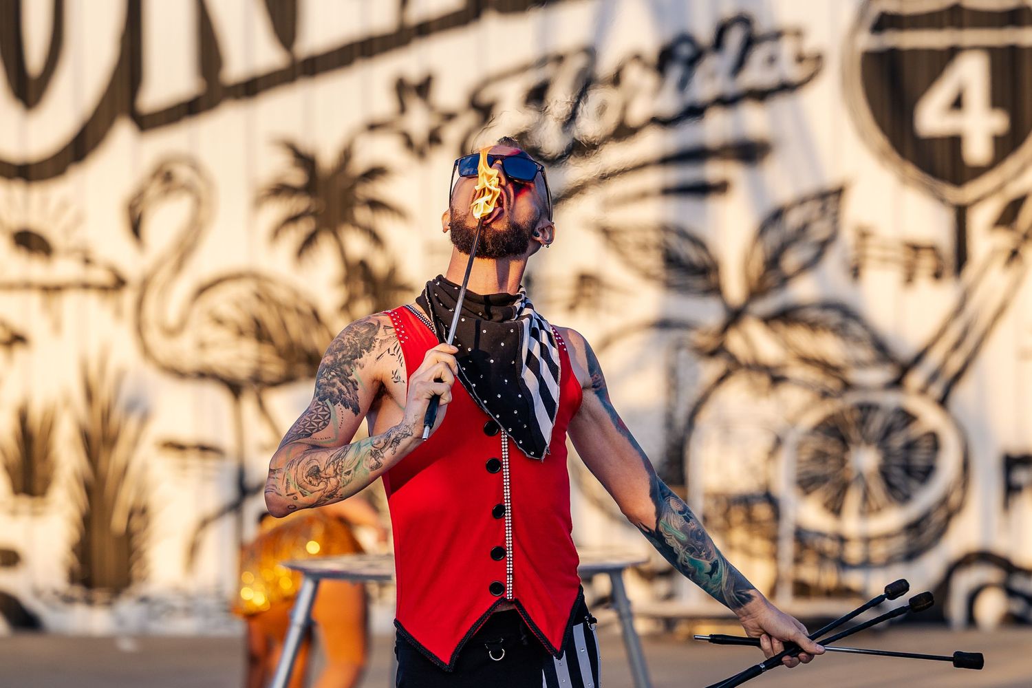 Tattooed circus performer in red vest and sunglasses performing a fire act at an outdoor Orlando event.
