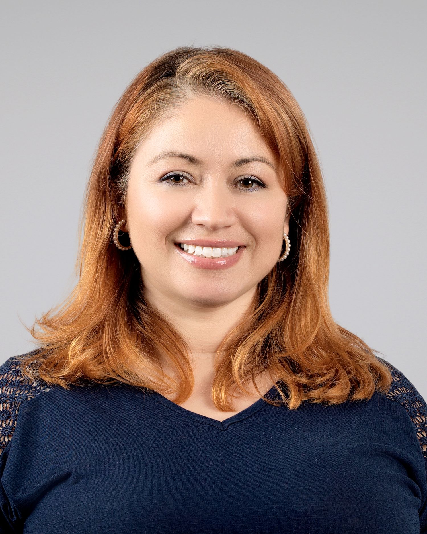 Smiling woman with auburn hair and gold earrings wearing a navy top poses for a professional headshot.